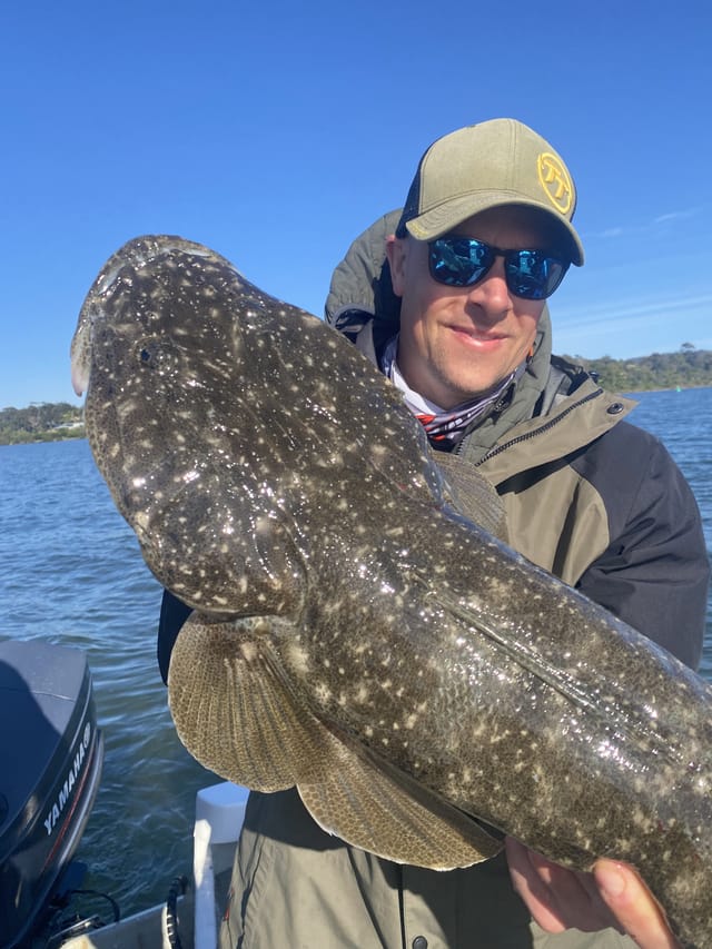 Adam holding a big dusky flathead across his chest on the boat, Mallacoota lake behind him