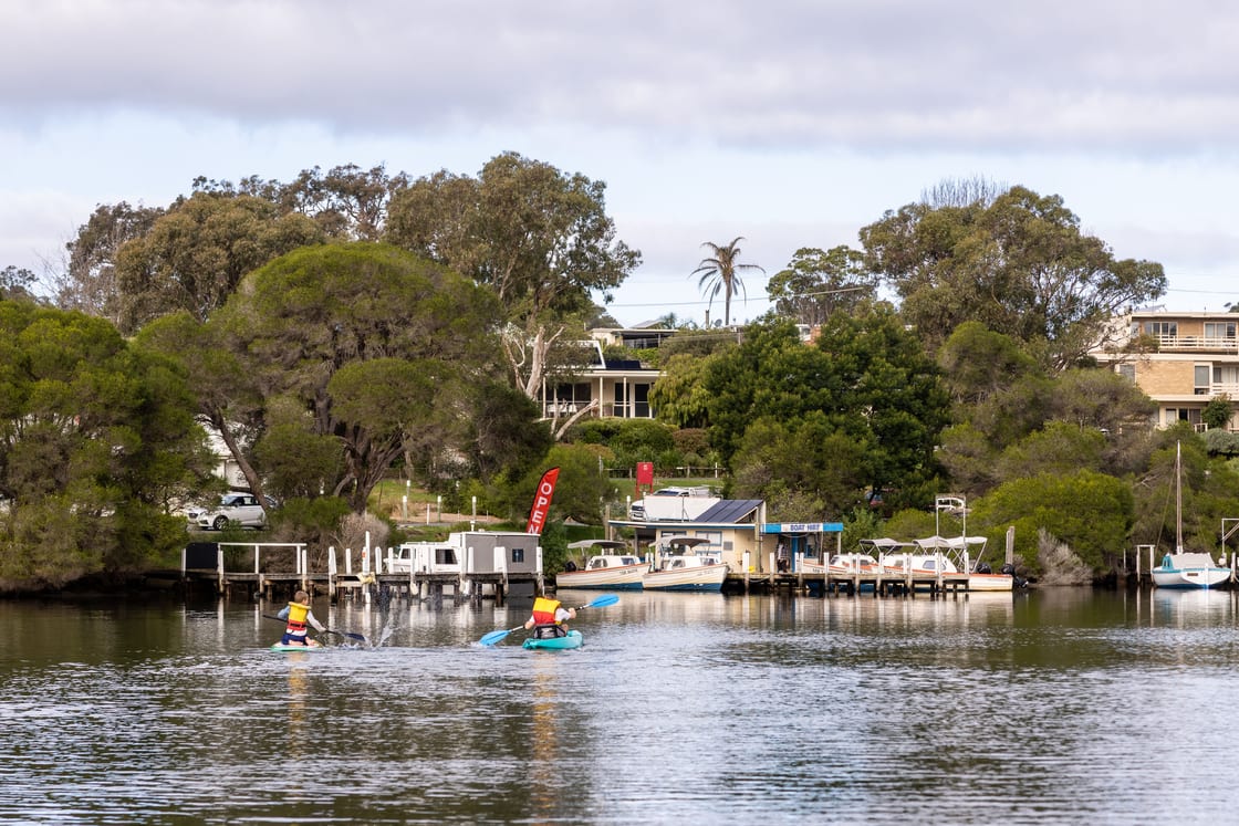 The Mallacoota Hireboats base seen across the water, with kayakers and the OPEN flag flying