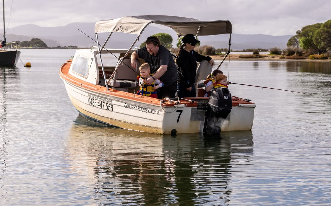 A father guiding his toddler's fishing rod aboard hire boat 7 with the family fishing off the stern, mountains behind