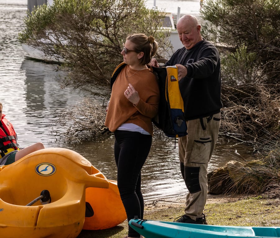 Grant helping a guest into her lifejacket at the water's edge, kayaks and pedal boats around them