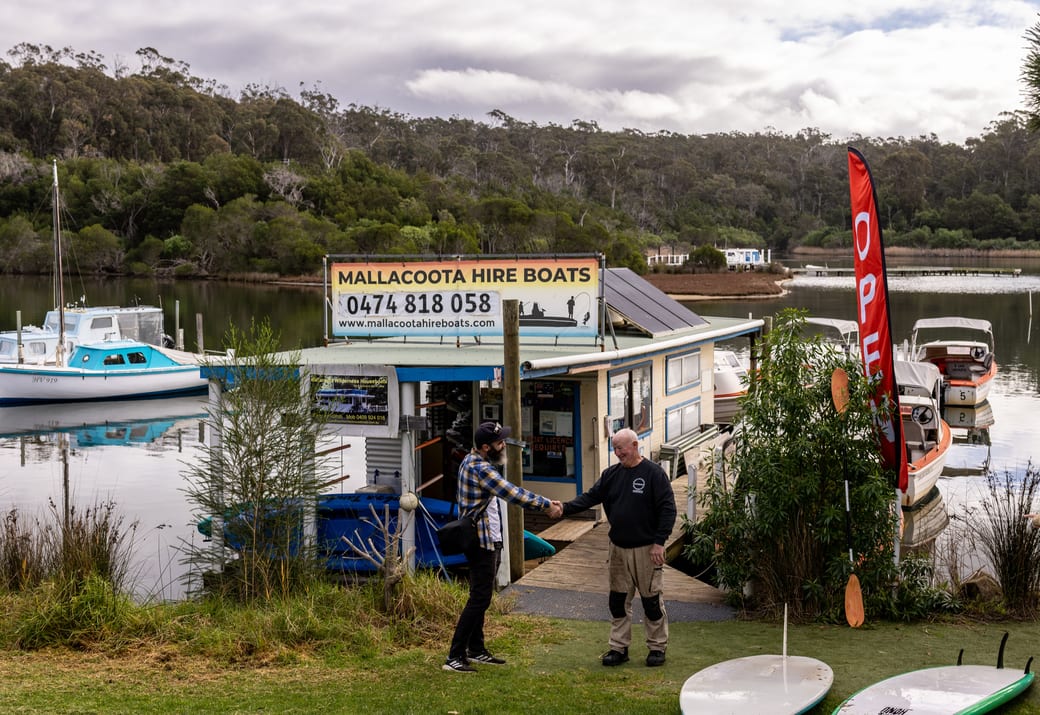 Grant welcoming a guest with a handshake at the kiosk, hand-painted sign and OPEN flag behind