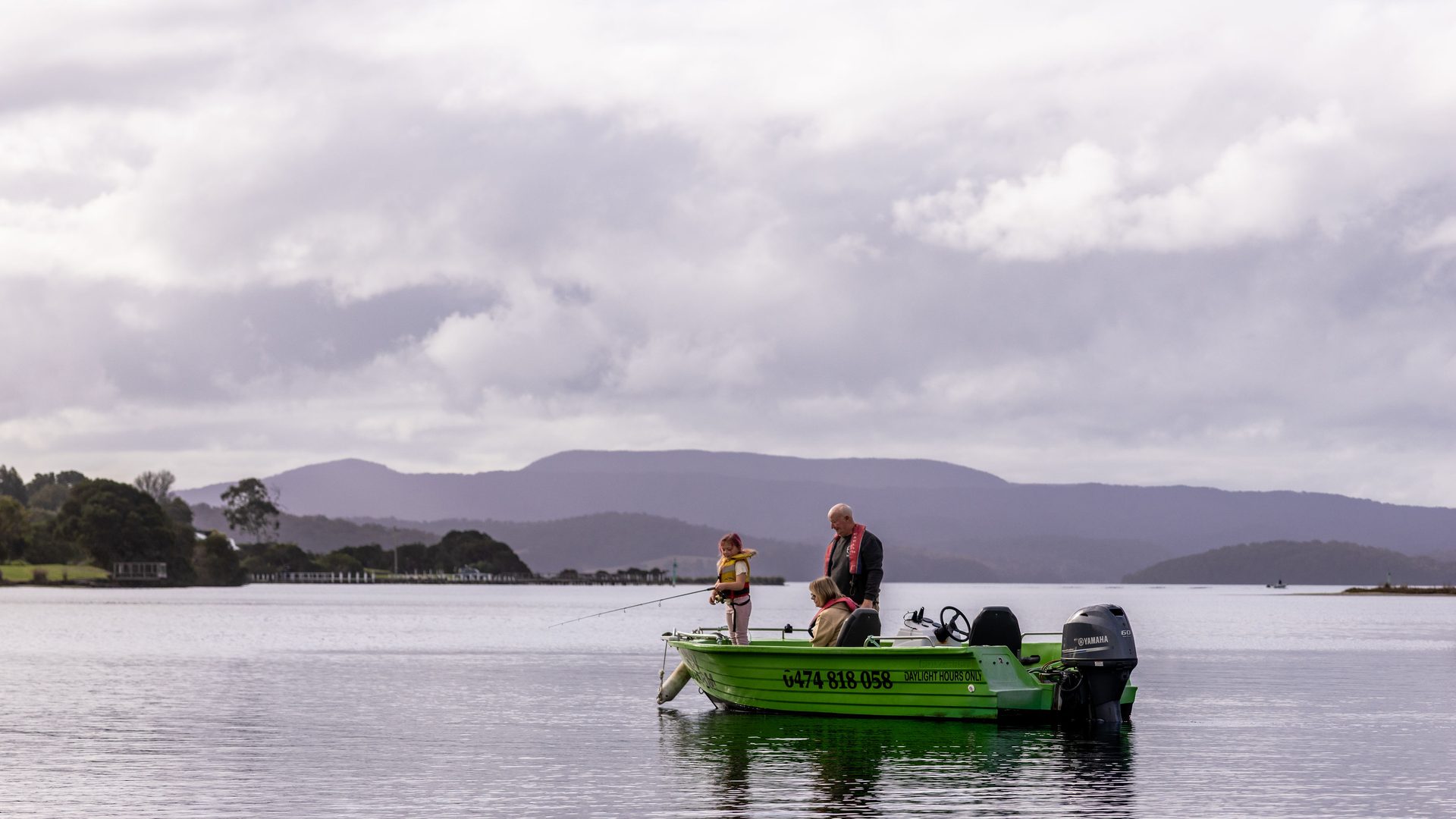 A family fishing at dusk from Kermit, the green hire boat, on the Mallacoota inlet with violet mountains behind