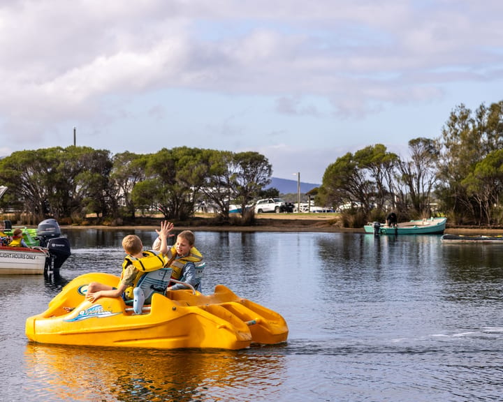 Two kids in yellow lifejackets waving from a yellow pedal boat