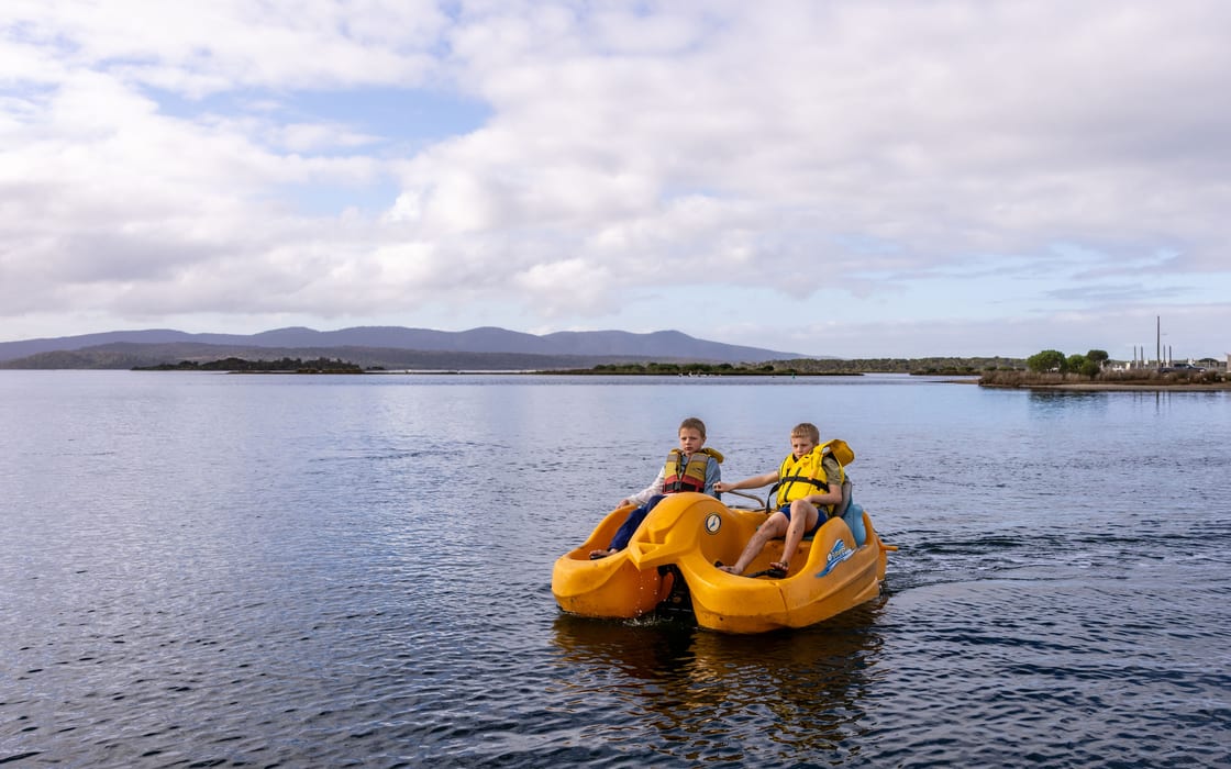 Two boys in yellow lifejackets cruising a pedal boat across calm open water