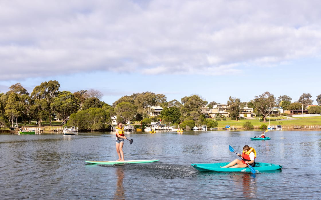 A stand-up paddleboarder and two kayakers in lifejackets on the inlet with Mallacoota township behind
