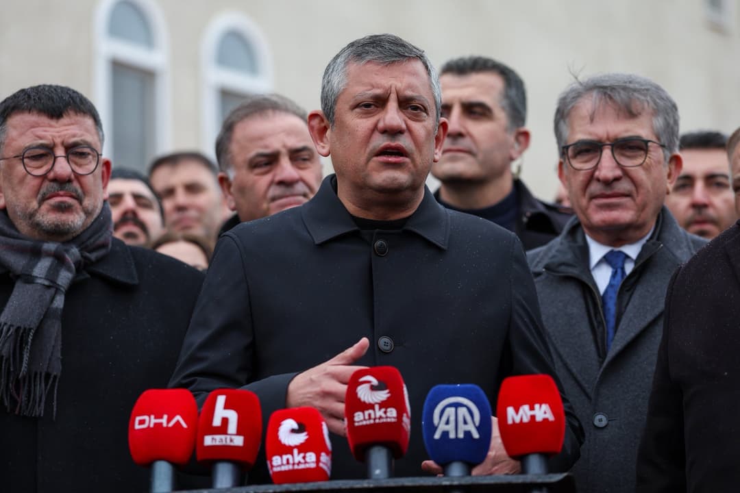 Açık havada, çok sayıda haber ajansı mikrofonunun önünde basın açıklaması yapan bir kişi; arkasında kalabalık bir grup duruyor.