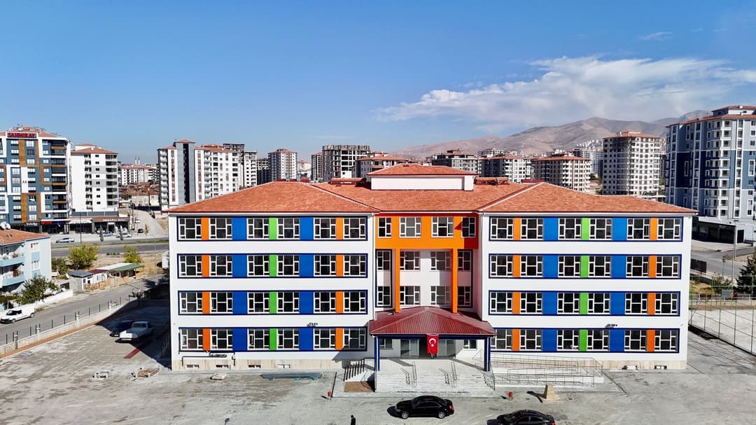 Havadan çekilmiş bir fotoğrafta, turuncu ve mavi cephe panelleriyle yeni yapılmış dört katlı bir okul binası; arkasında toplu konutlar ve dağlar görünüyor.