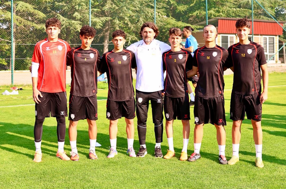 Antrenman sahasında yan yana dizilmiş altı genç futbolcu ve aralarında beyaz eşofmanlı bir antrenör poz veriyor.