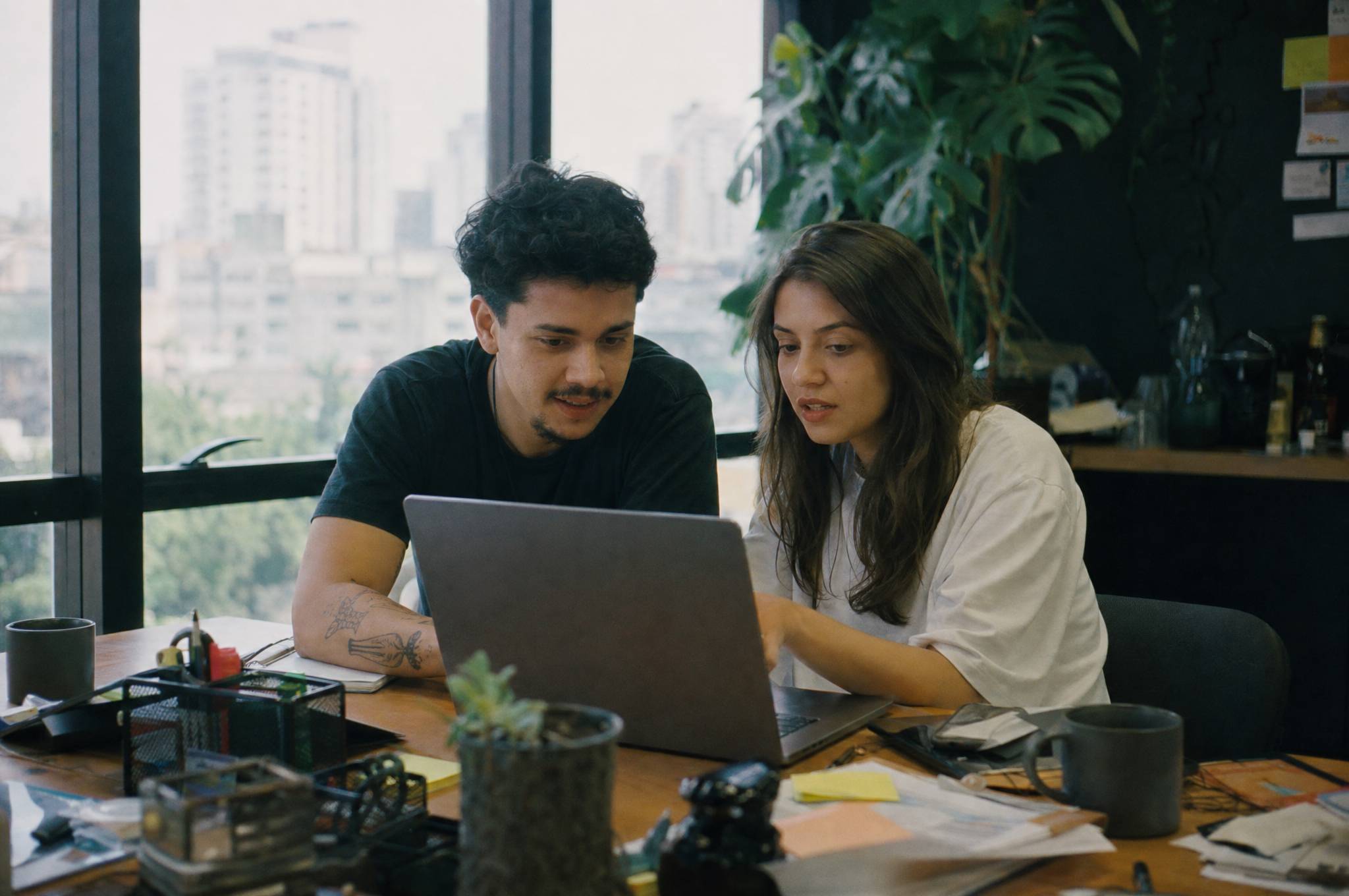 Dois gestores de tráfego conferindo o mesmo notebook numa mesa de agência, com a cidade na janela