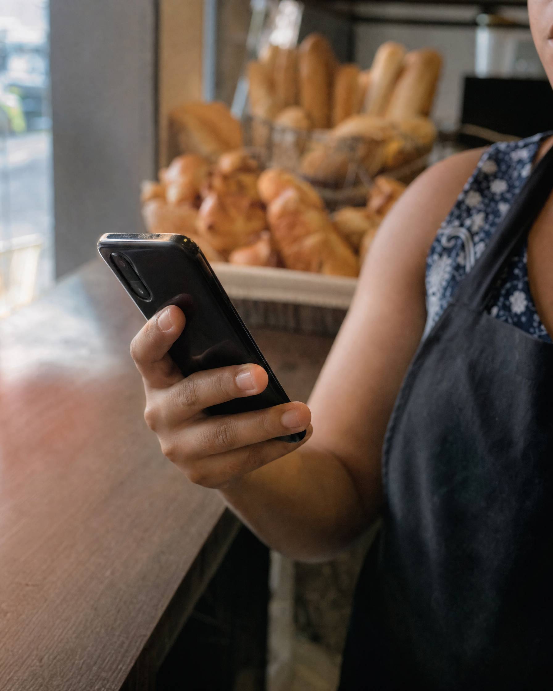 Mão de uma dona de padaria segurando o celular no balcão, com cestas de pão ao fundo