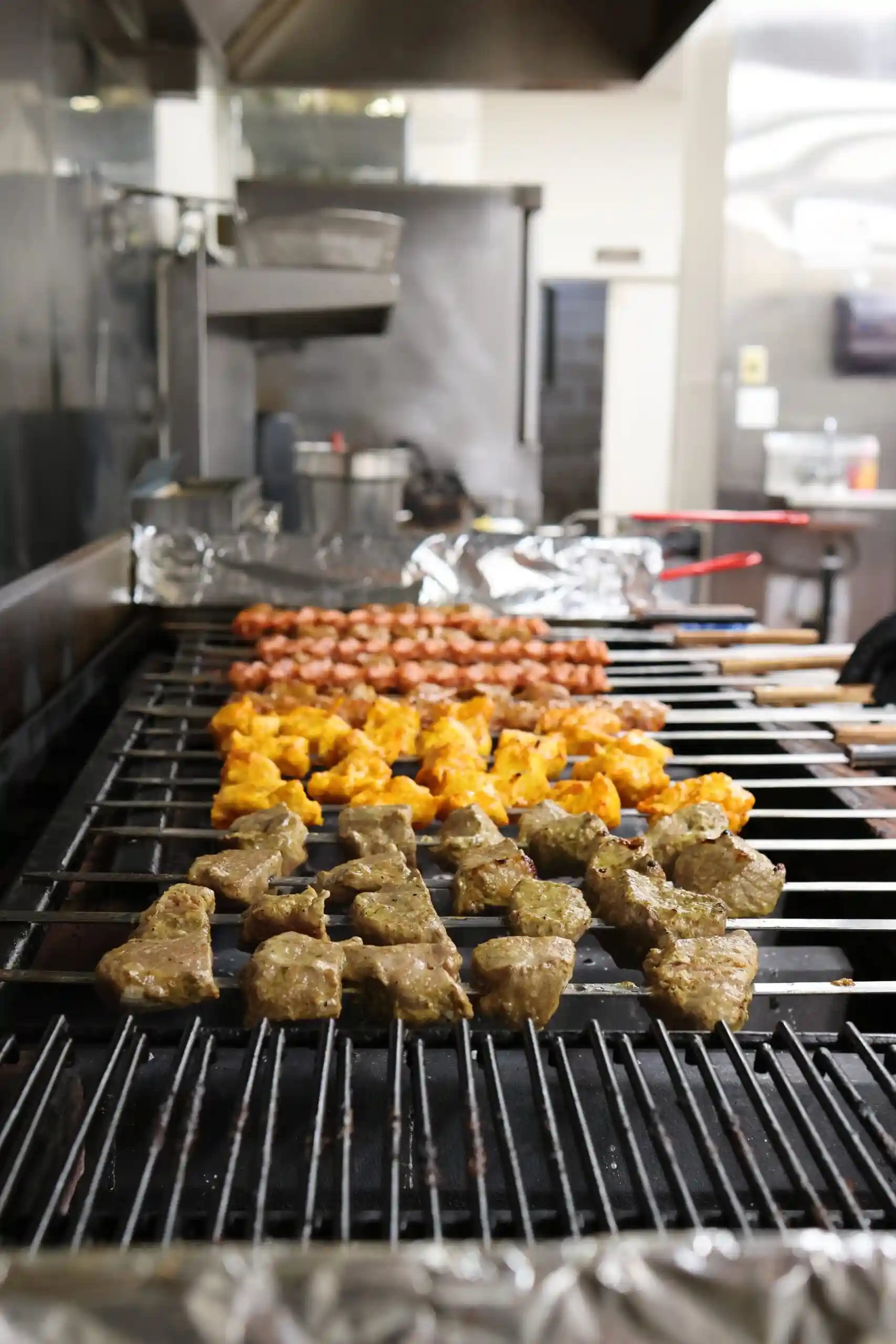 Grilled beef, chicken, and sausage skewers cooking on an open flame inside a professional kitchen