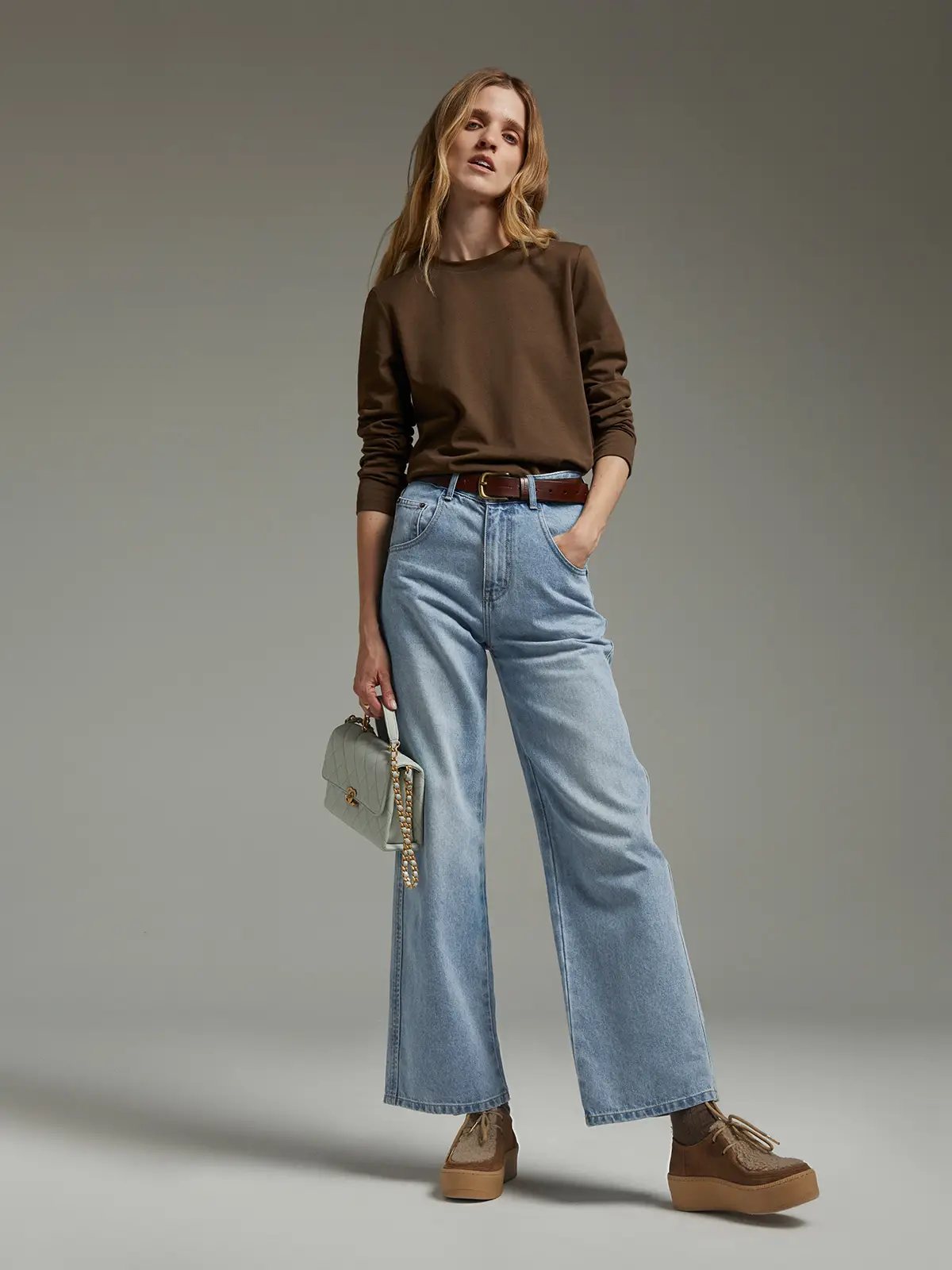 Model wearing a brown long-sleeve crewneck T-shirt tucked into light-wash wide-leg jeans with a brown belt and holding a small light-colored bag.