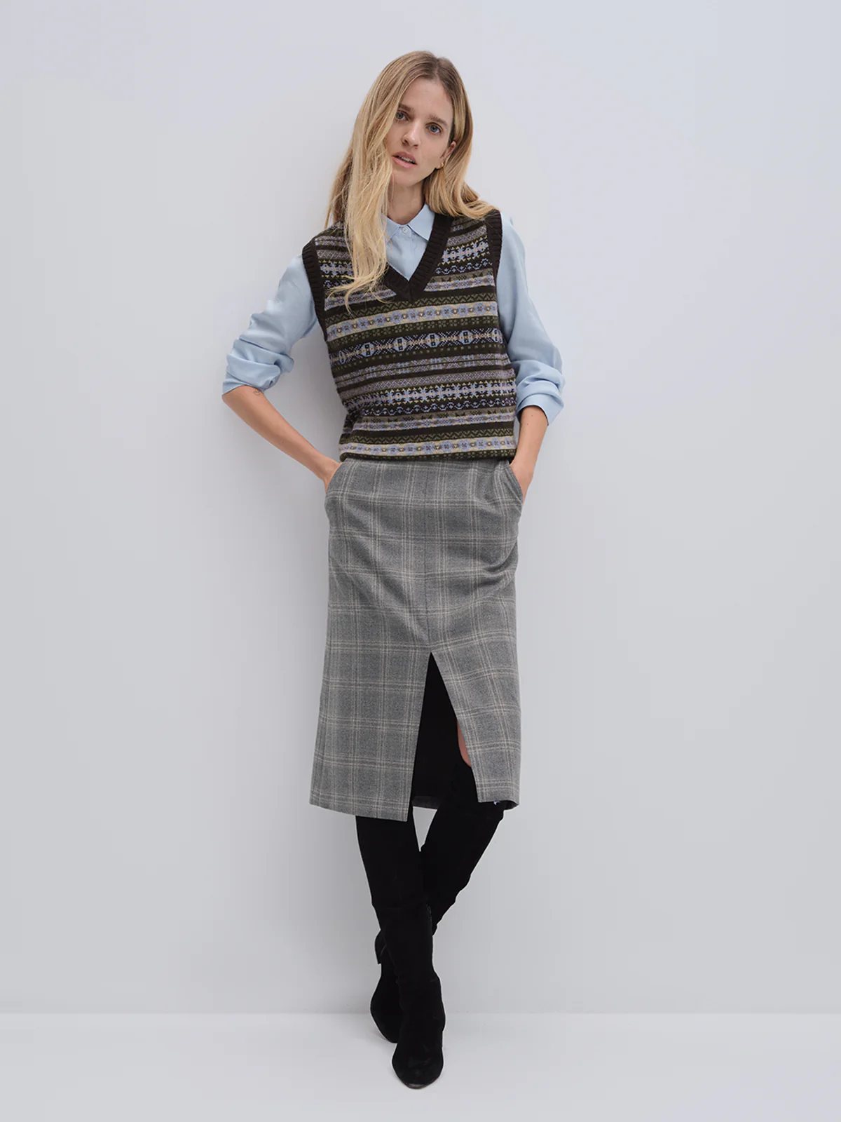 Front view of model in andreatang blue-grey wool check midi skirt with patterned vest and boots