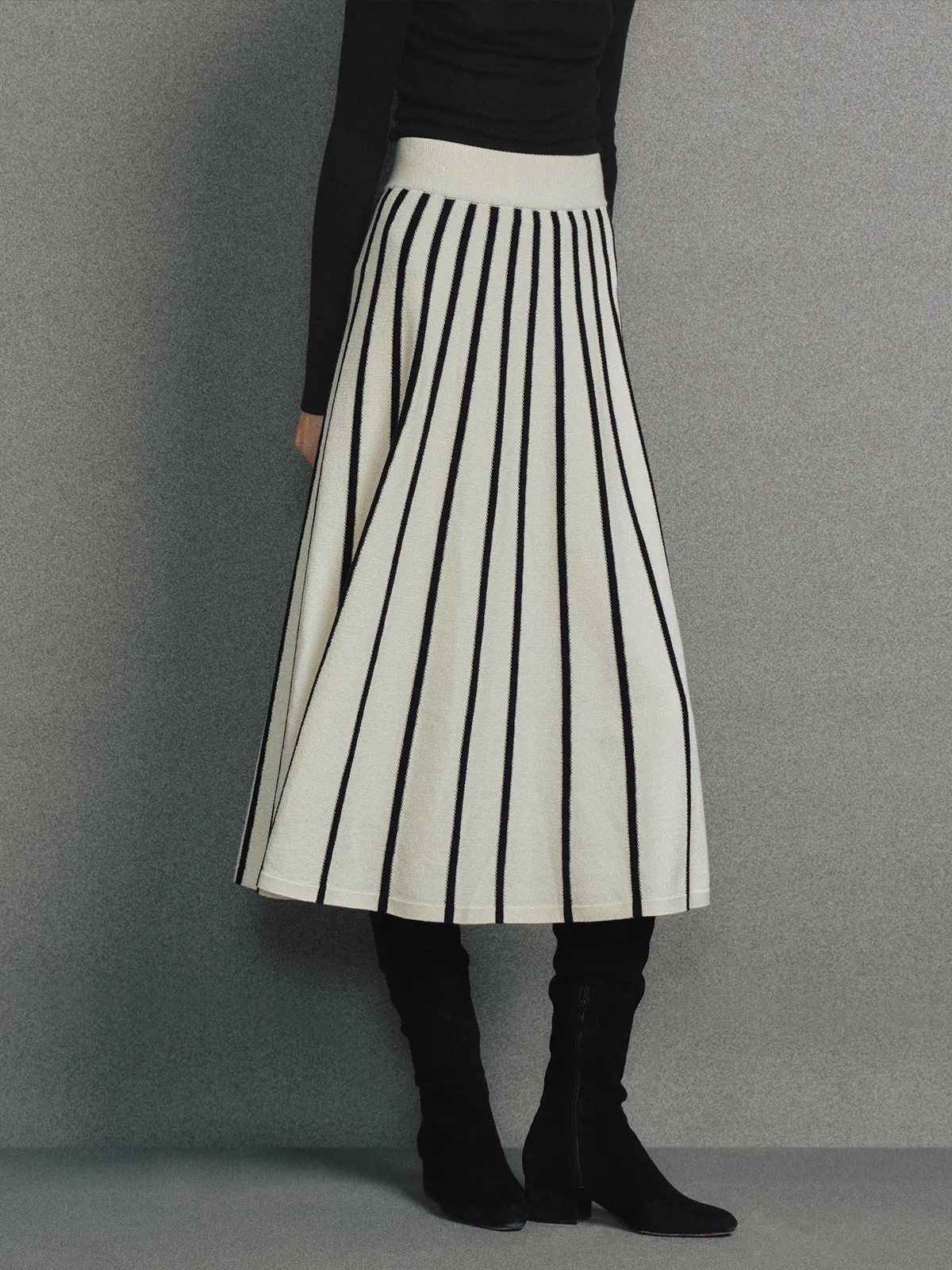 Side view of a model in a Black/White vertical stripe A-line knit skirt with white waistband, black top, and black boots.