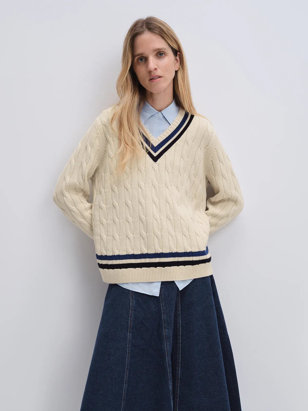 Off-white cable knit V-neck tennis jumper with contrasting trim, worn over a turtleneck in a relaxed side pose.