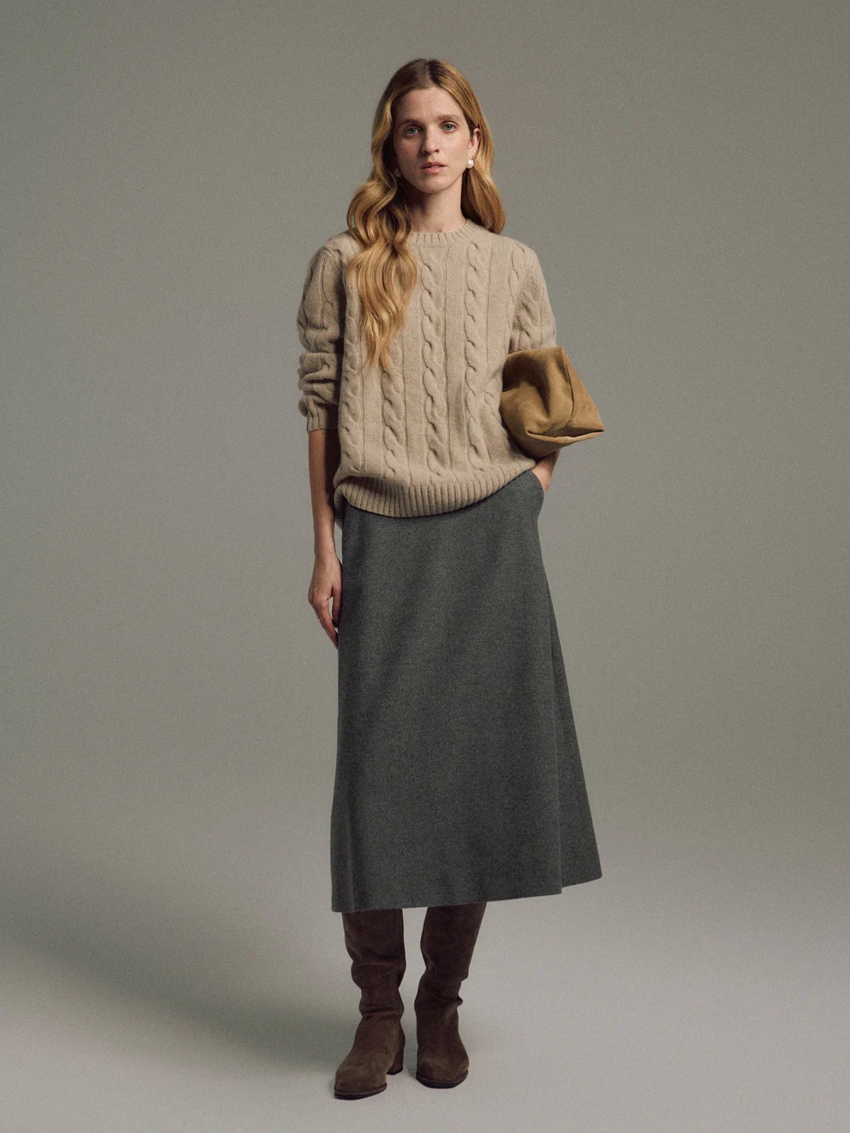 Model wearing an apricot cable knit jumper layered over a shirt, paired with a midi skirt and boots, holding a clutch.