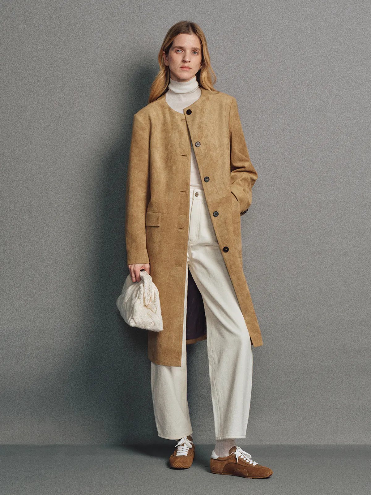 Side view of model in a camel faux suede collarless long coat with front buttons, holding a matching camel bag, over trousers.