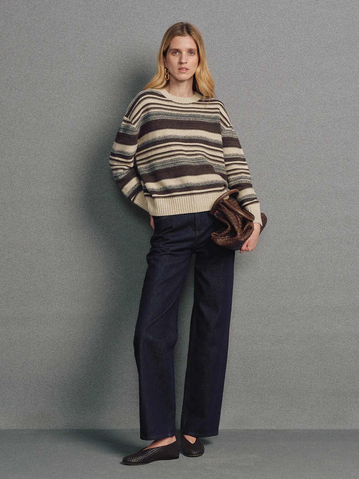 Front view of model wearing a brown irregular multicolour stripe wool cotton jumper holding a bag.