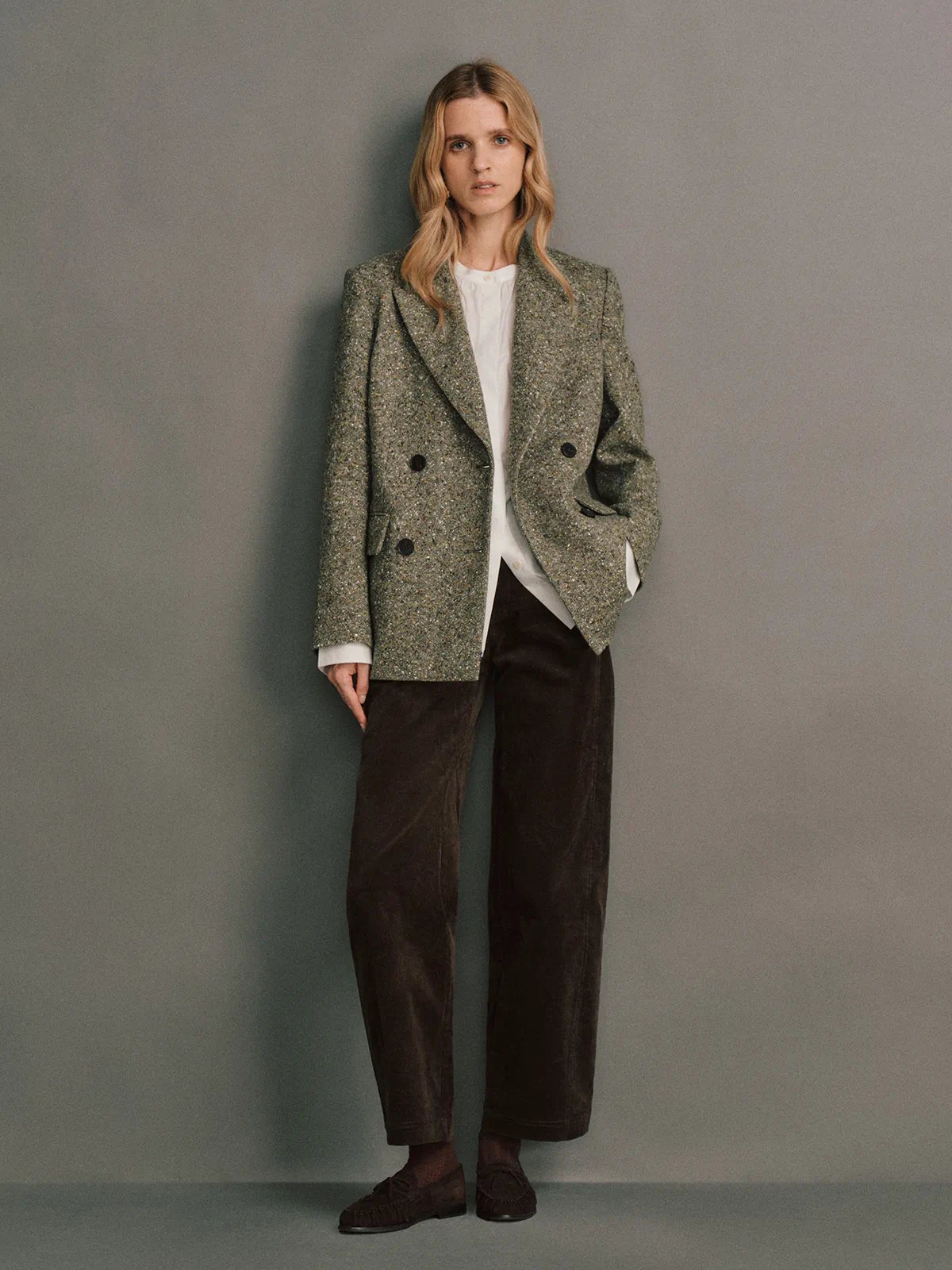 Front view of a model wearing a green wool flecked tweed double-breasted blazer with flap pockets and dark buttons.