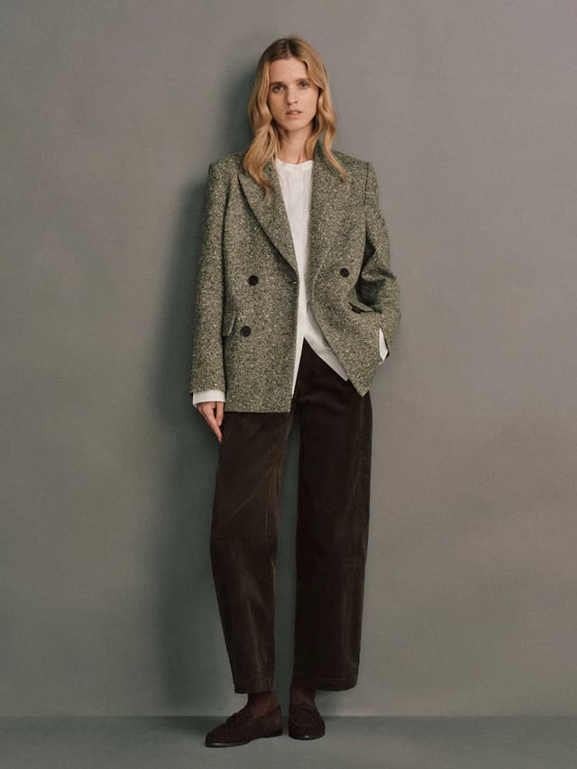 Front view of a model wearing a green wool flecked tweed double-breasted blazer with flap pockets and dark buttons.