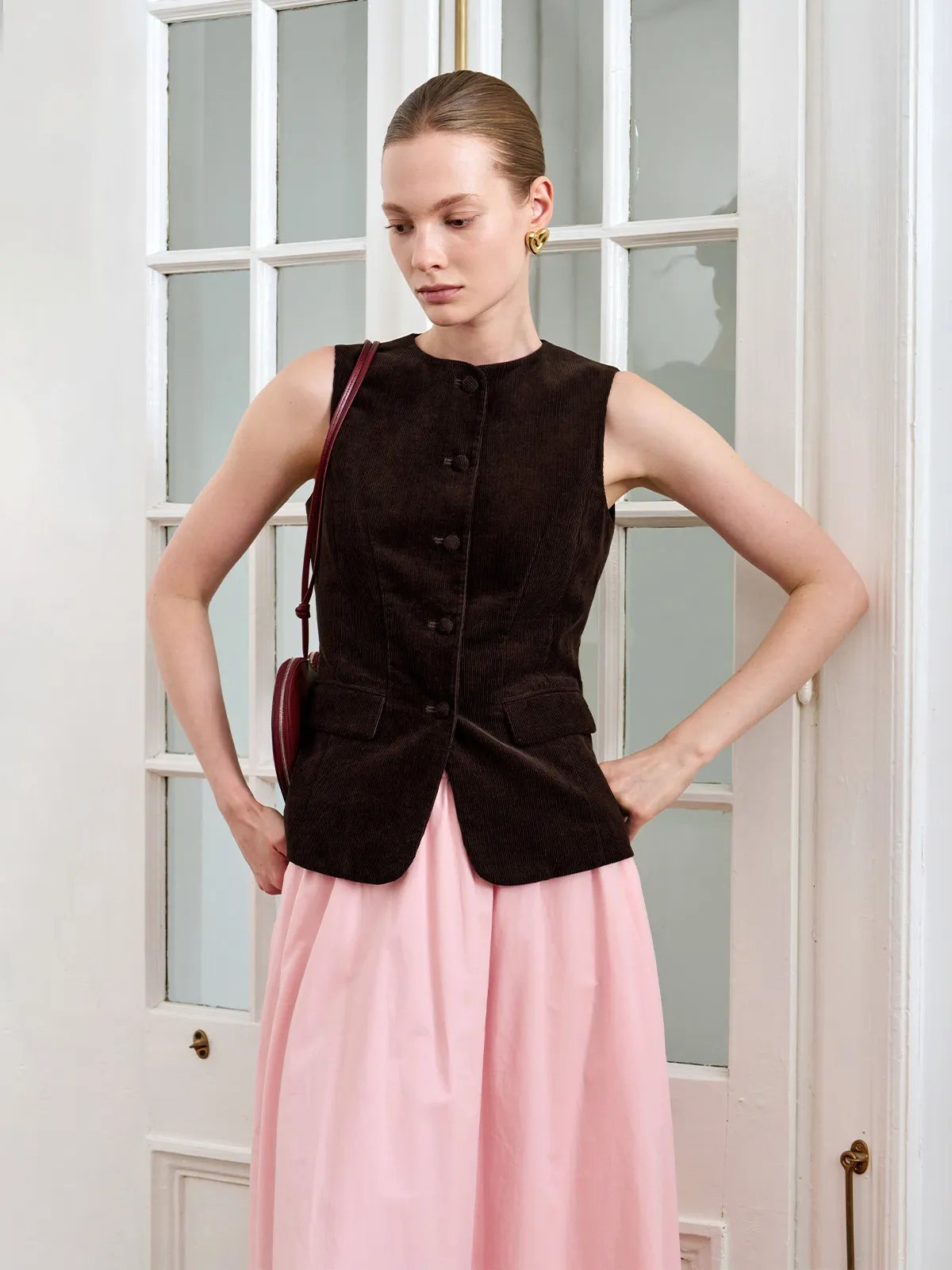 AIDAN vest styled with pink skirt and burgundy circular crossbody bag, model leaning against door