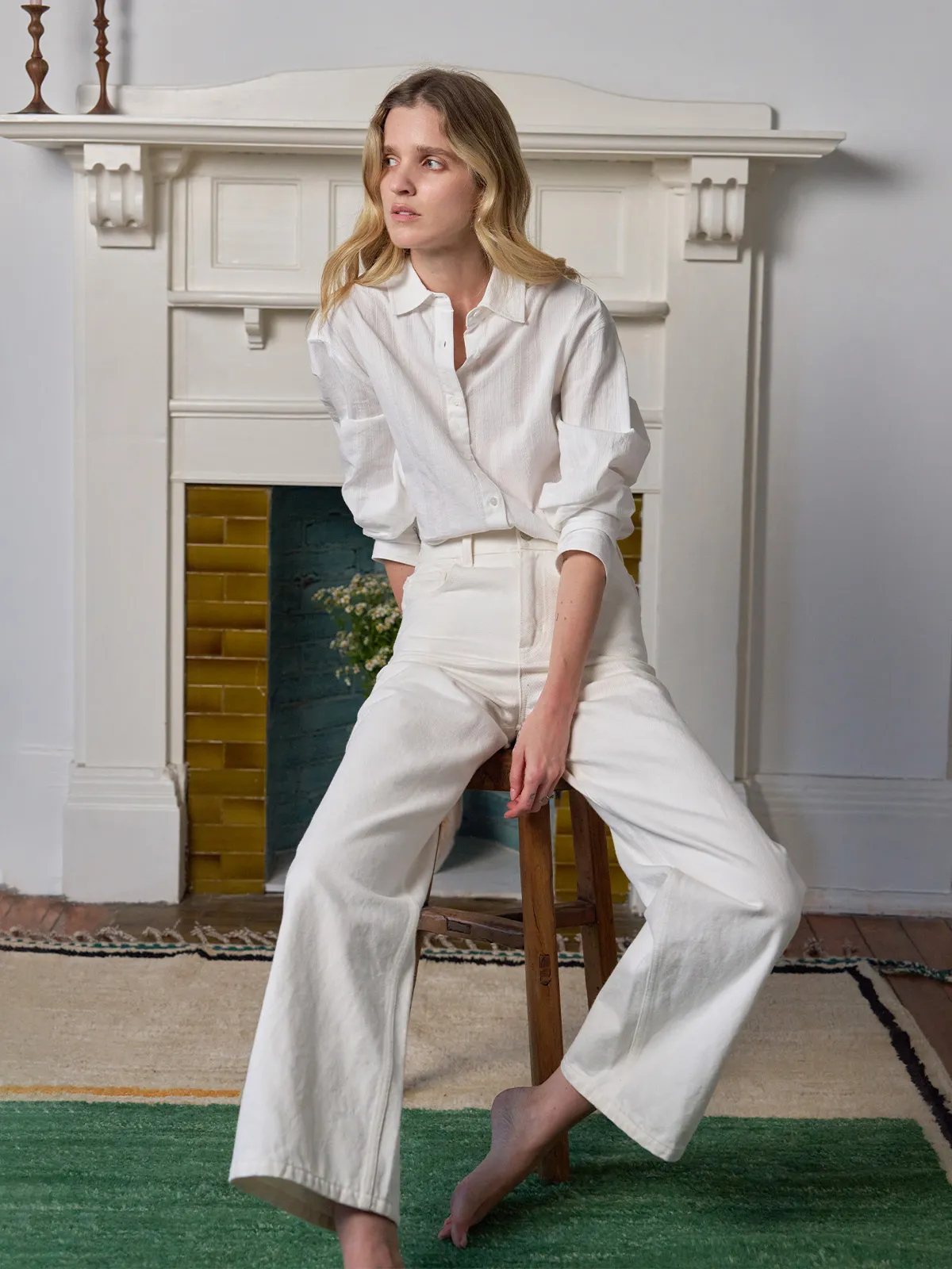 Model seated sideways in RORY shirt and white wide-leg trousers near fireplace