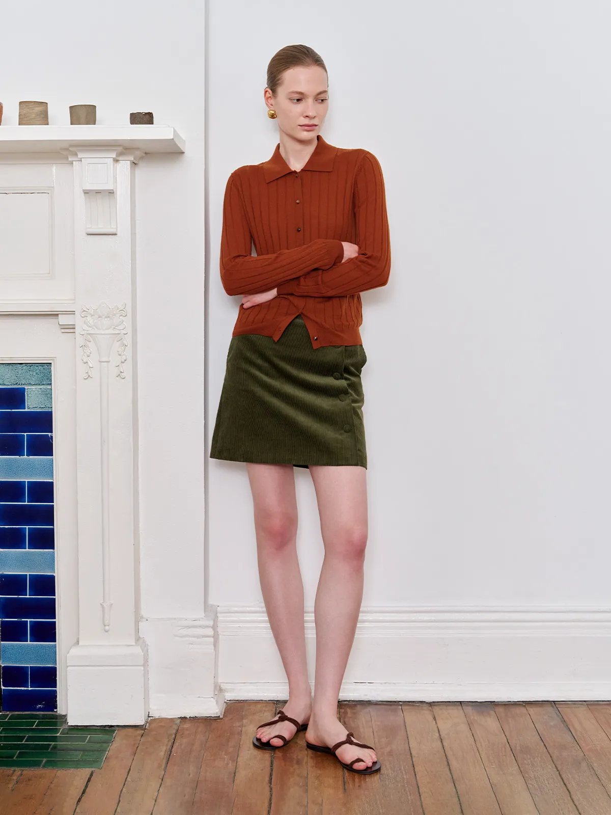Model in terracotta cardigan with arms crossed, wearing olive mini skirt against white wall and fireplace