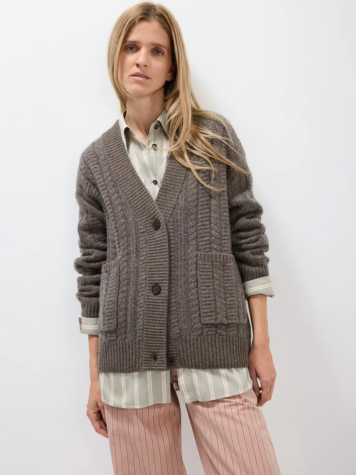 Model wearing a coffee-colored V-neck cable knit cardigan with front buttons and patch pockets over a striped shirt and pants.
