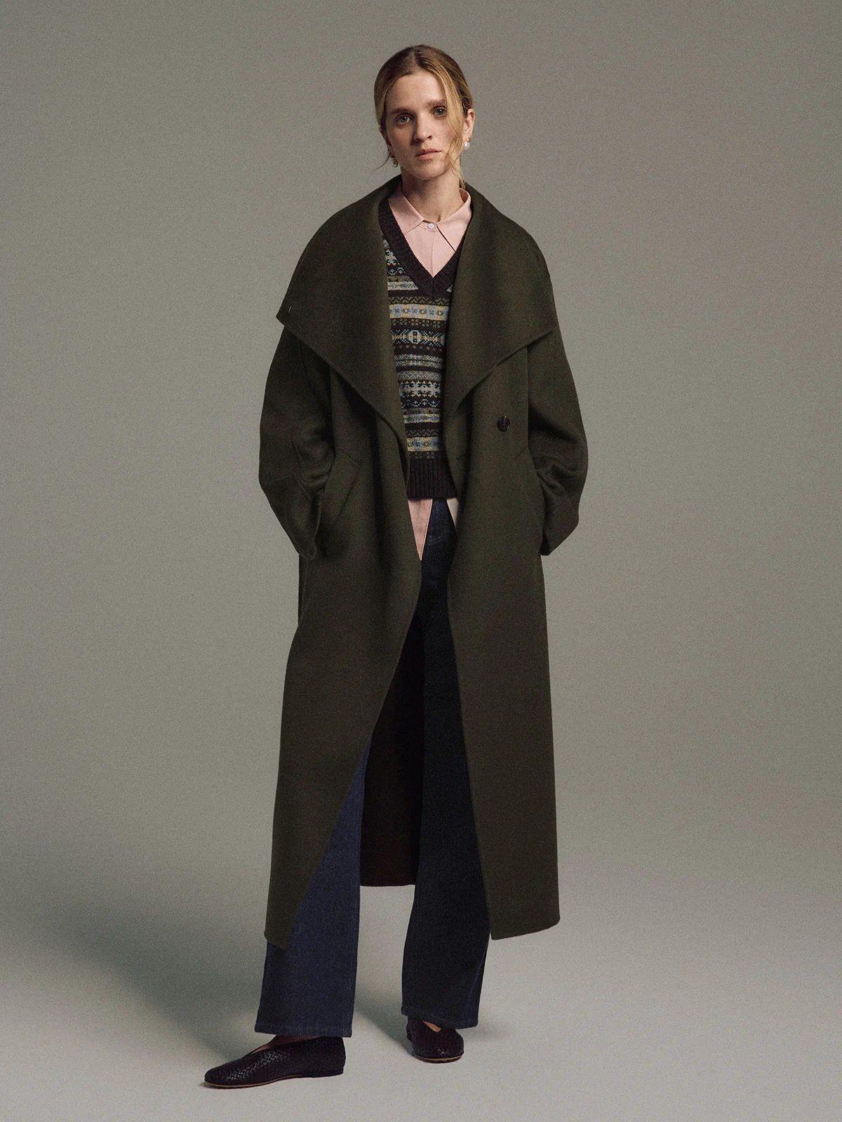 Model in the Olive TANSY long coat over a patterned sweater and dark trousers, hands in pockets, front-facing.