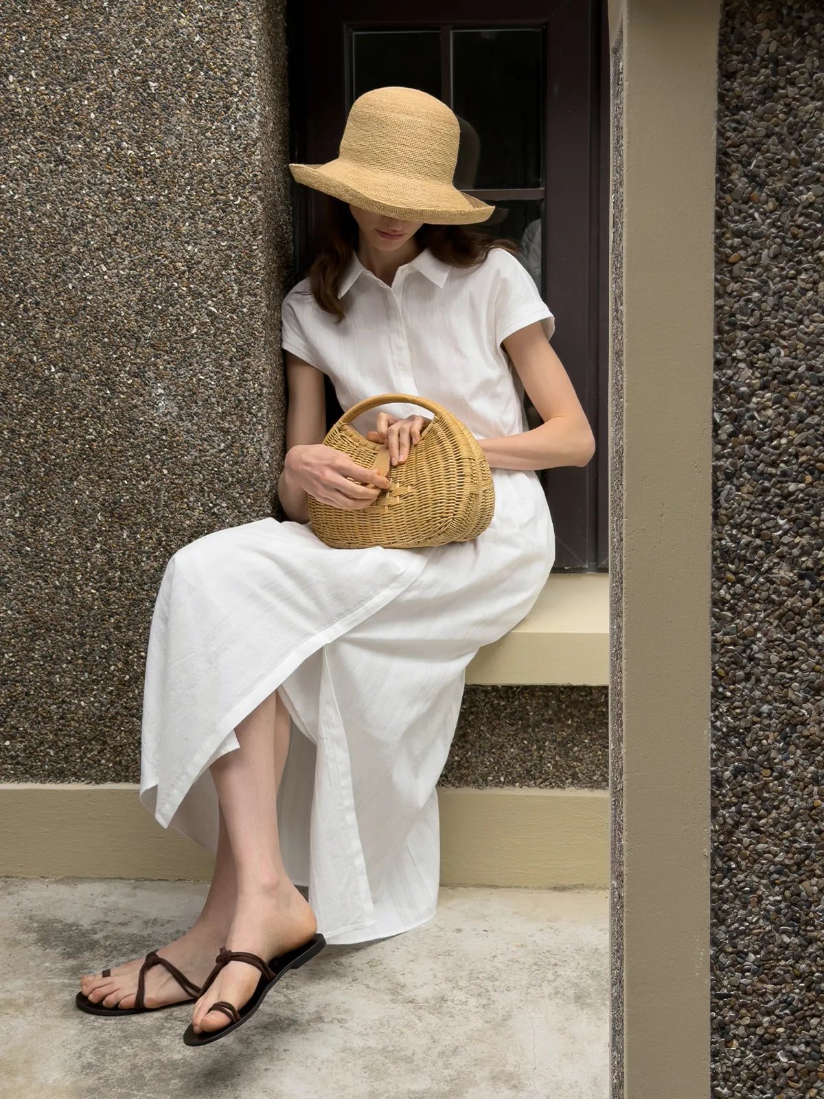 Woman seated outdoors in white shirt dress, straw hat, and woven bag