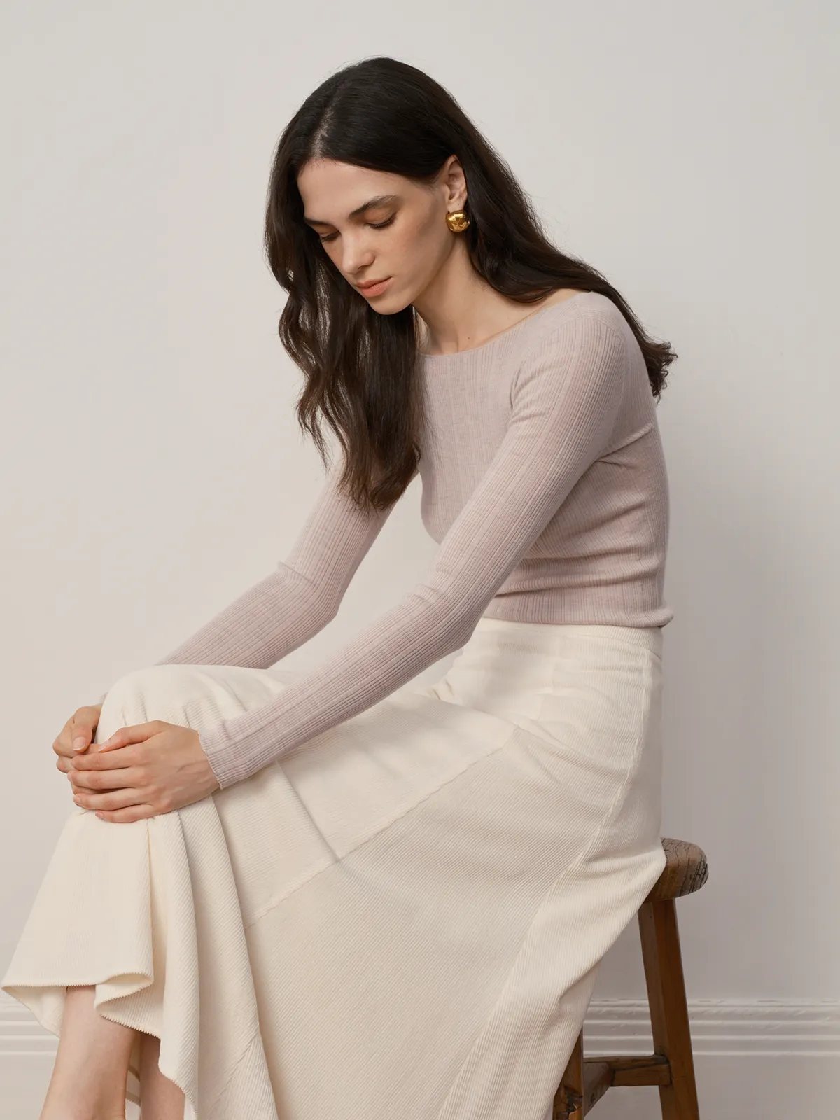 Model seated on wooden stool wearing THERESA knit top and cream asymmetrical skirt