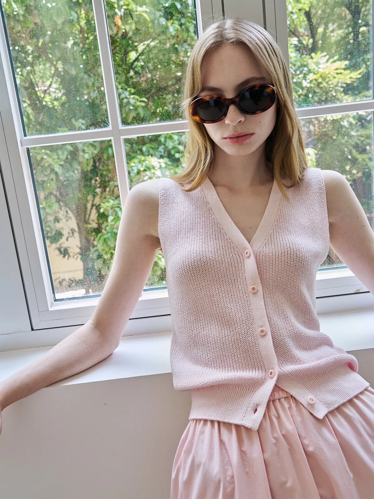 Model wearing blush ZOLA vest and pink skirt, leaning on windowsill with sunglasses