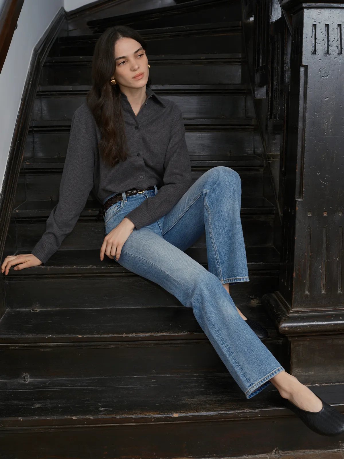 Model seated on black wooden stairs wearing JOSETTE jeans with gray cardigan and braided belt