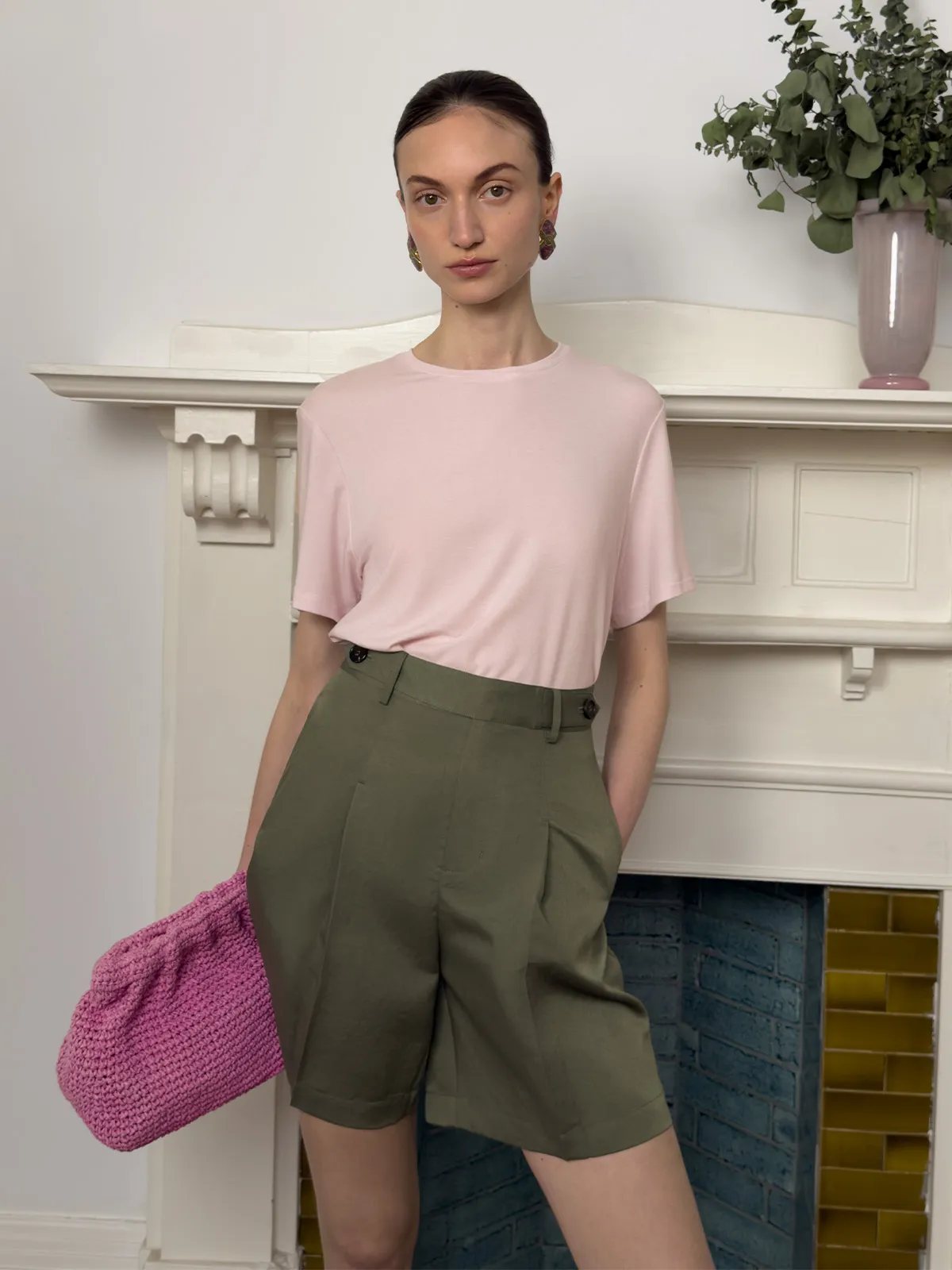 Model in olive green ADELE Bermuda shorts with pink tee and pink crocheted clutch at fireplace