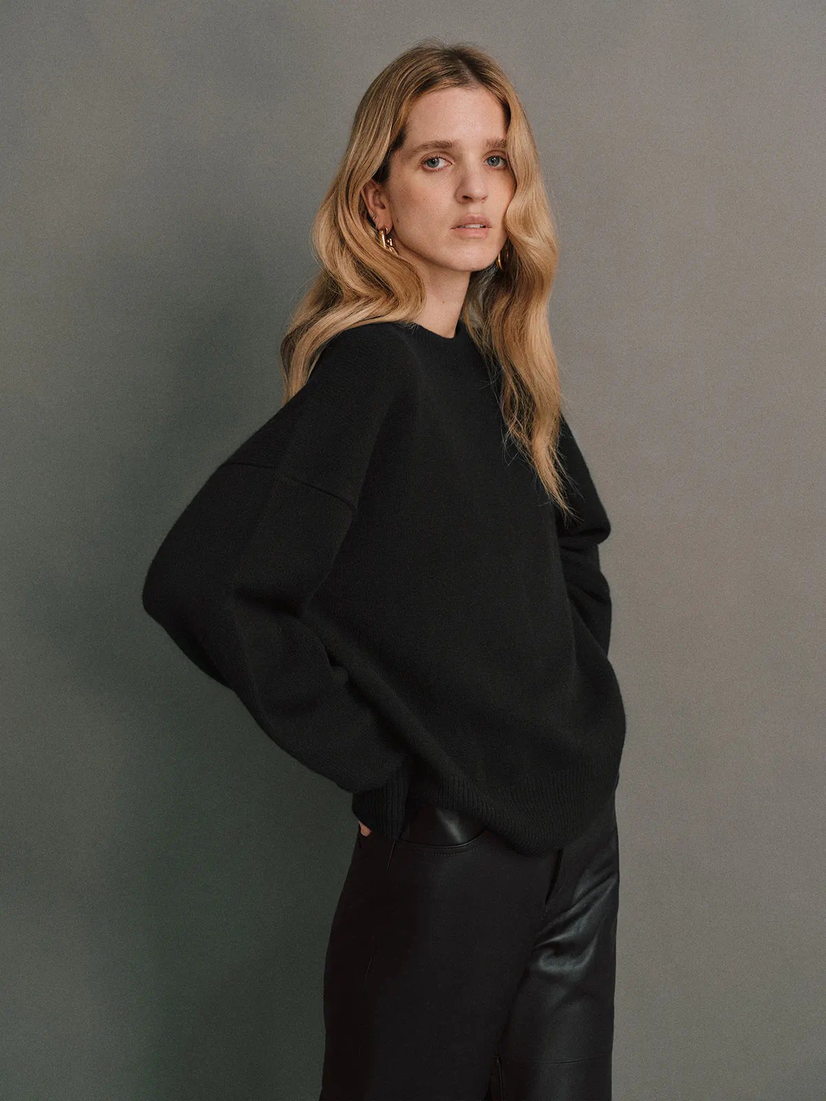 Side view of model in a Black merino wool crewneck jumper with dropped shoulders, styled with black leather trousers.