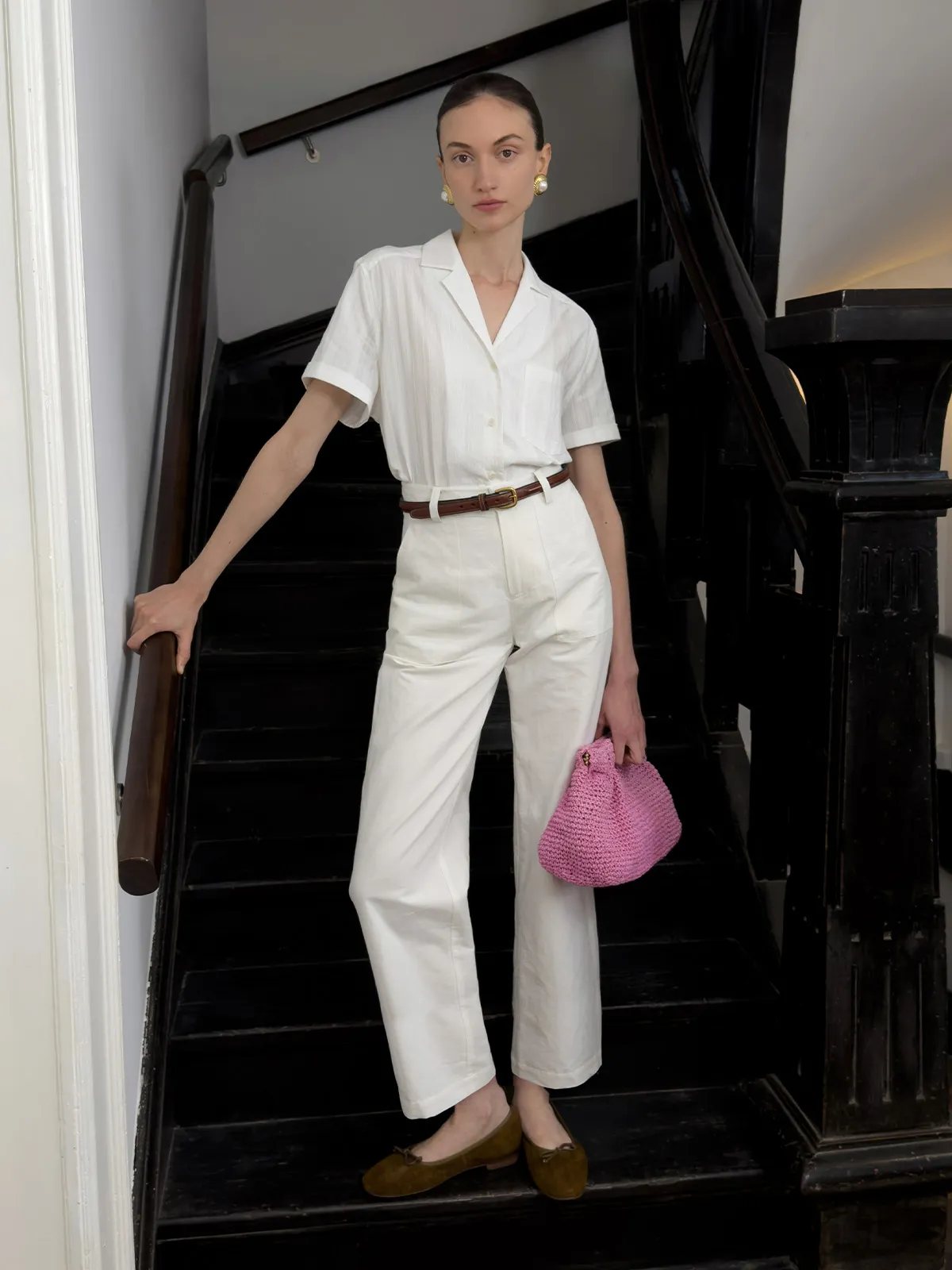 Model descending black staircase in white REID cropped trousers, ivory shirt, brown belt, and pink crocheted bag