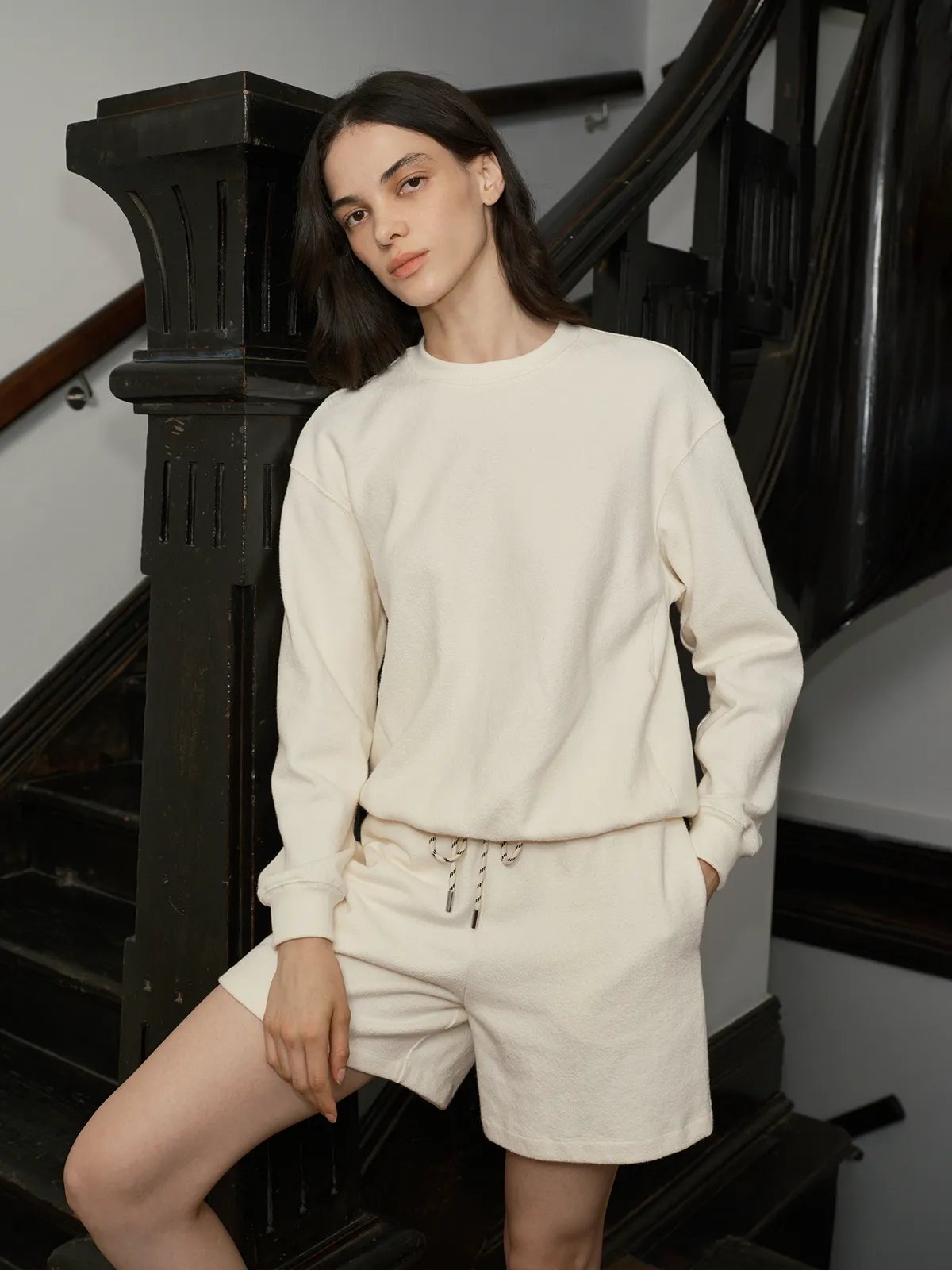 Model in ivory ARLO terry short sweatpants with matching crewneck, leaning on black staircase banister
