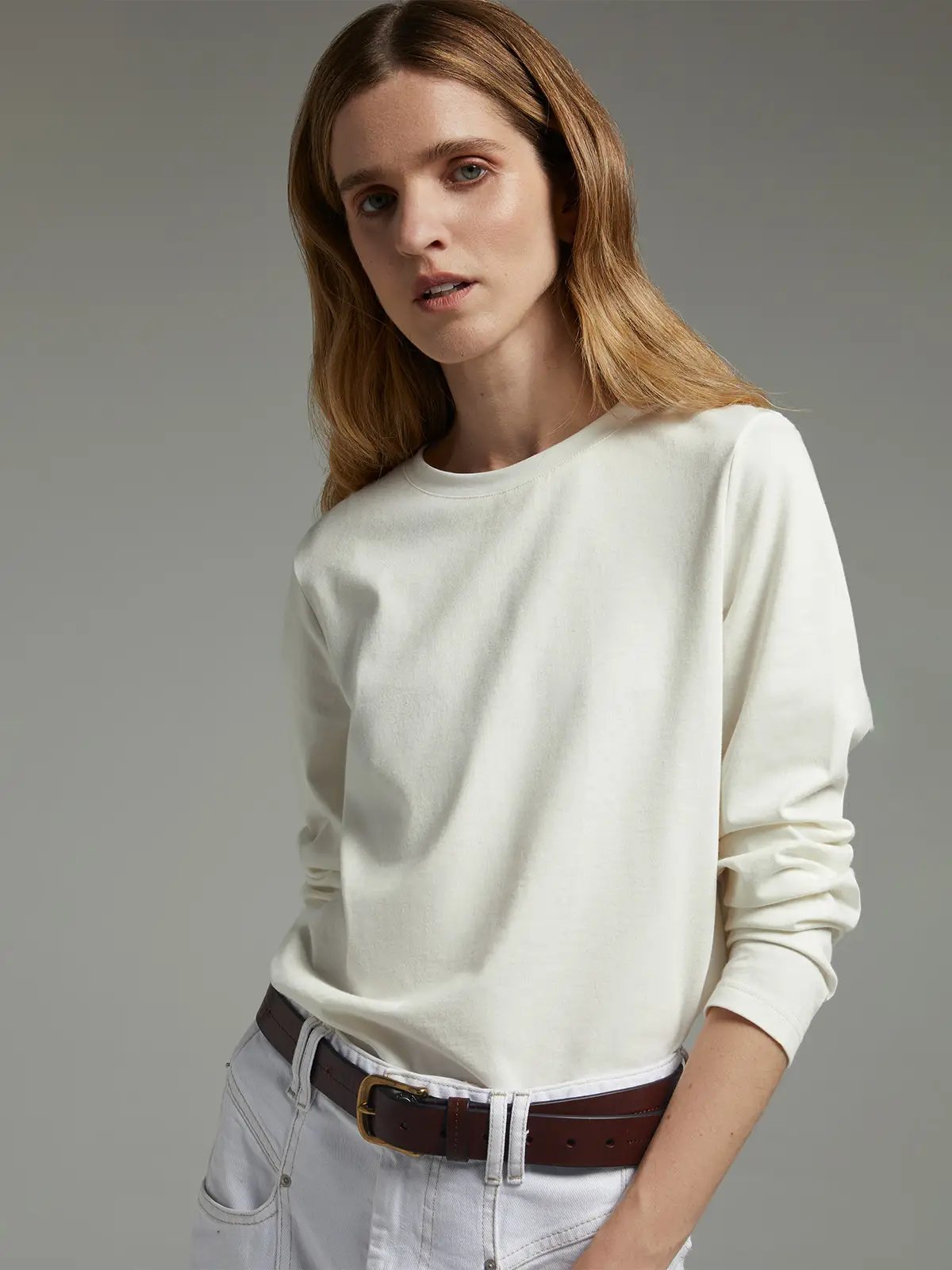 Model wearing a white long-sleeve crewneck T-shirt with rolled sleeves, tucked into light-wash jeans with a belt.