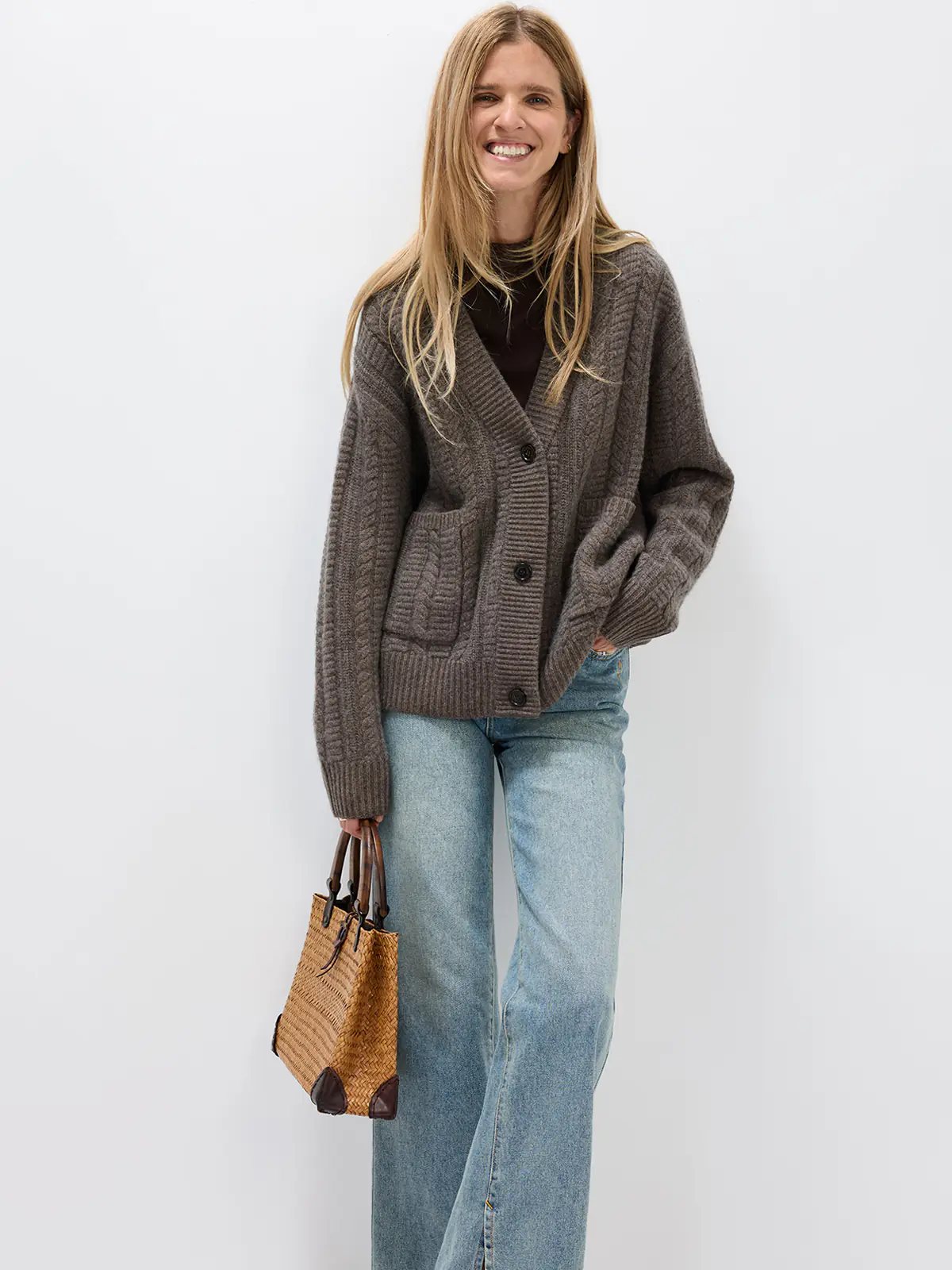 Model wearing a green V-neck cable knit cardigan with front buttons and patch pockets over a plaid shirt and skirt with knee-high boots.