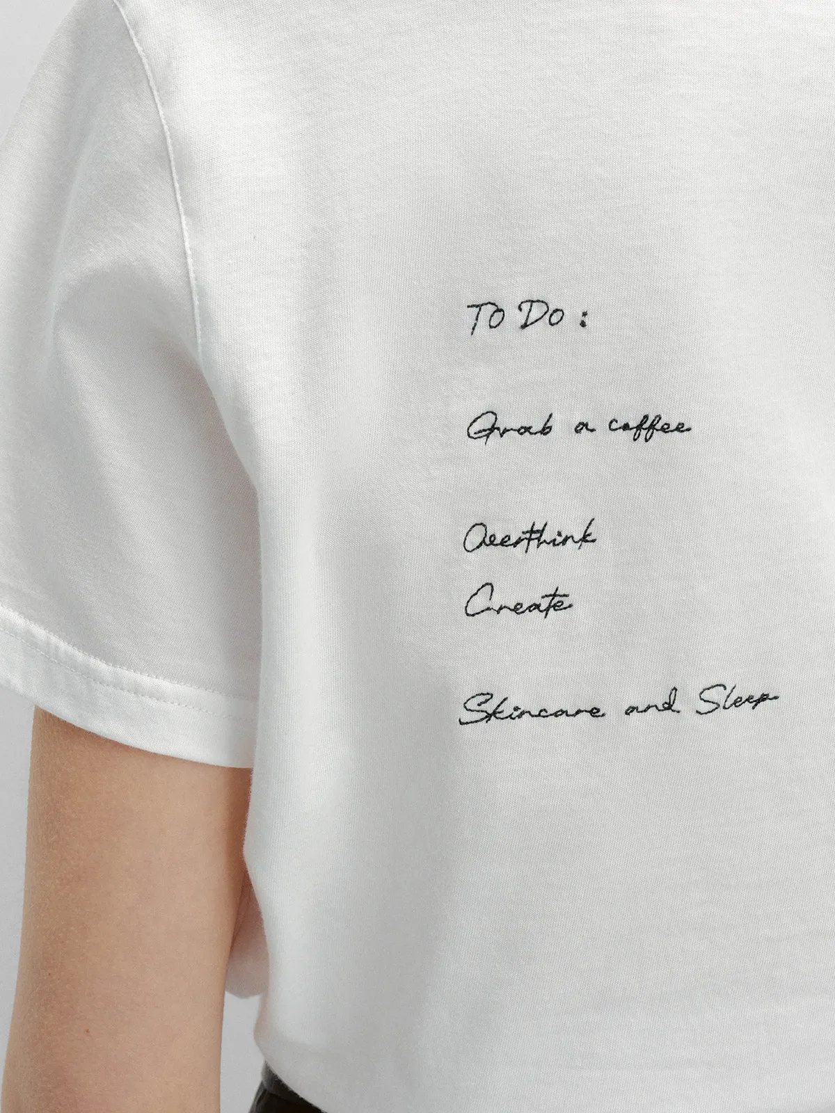 Close-up of black hand-embroidered text on back of white t-shirt: 'To Do: Grab a coffee, Overthink, Create, Skincare and Sleep'