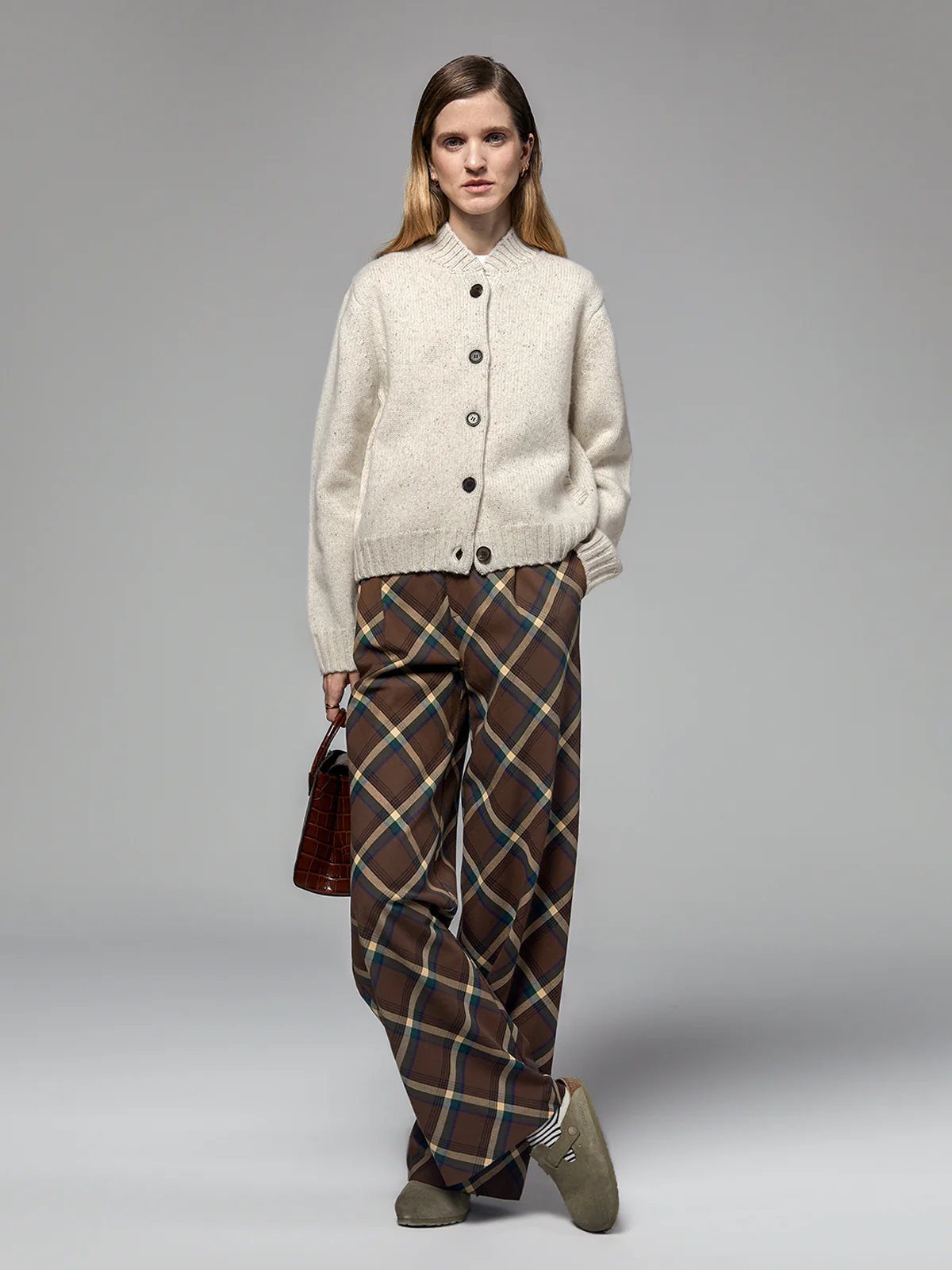 Model wearing the Oatmeal Merino Wool Baseball Cardigan open over a shirt and trousers, holding a bag.
