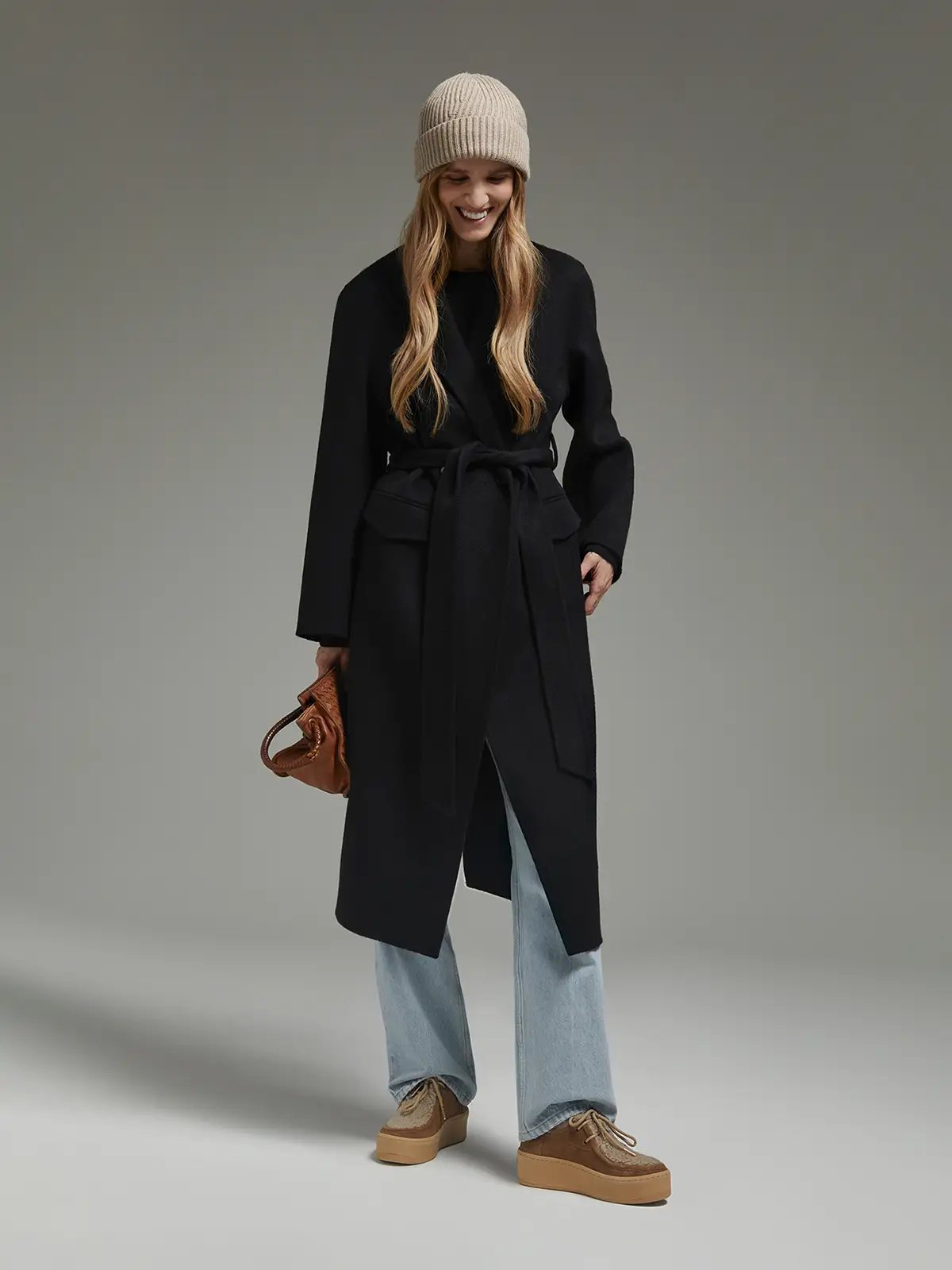 Front view of a model in a long belted coat, light jeans, and platform shoes.