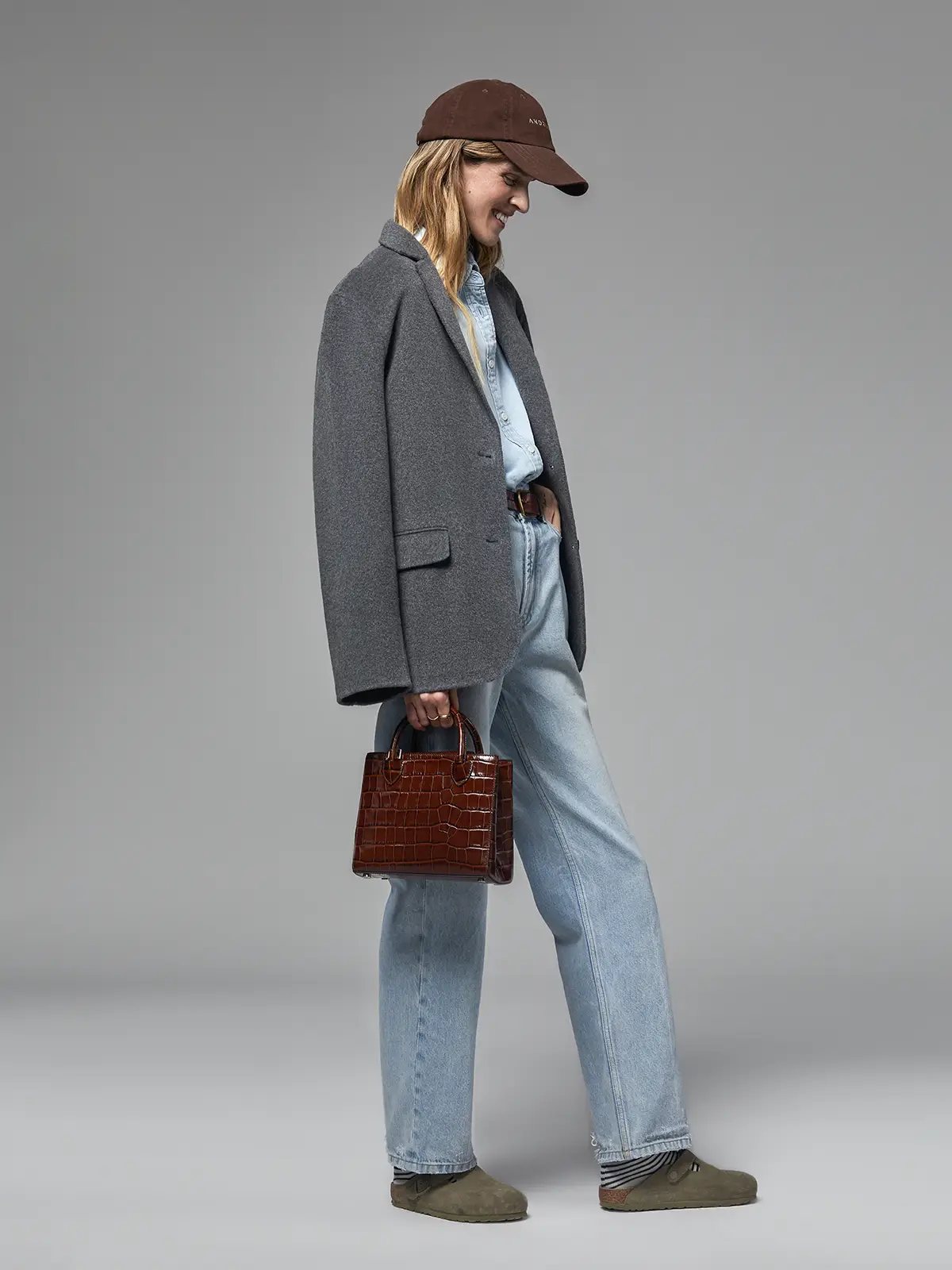 Side view of model in a Mixed Gray merino wool blazer, light shirt, jeans, cap, and holding a bag.
