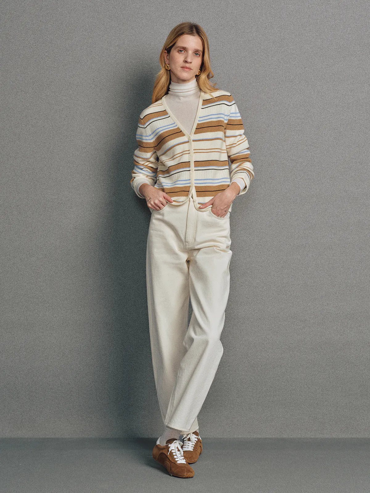 Model wearing the Camel multicolour stripe V-neck cardigan layered over a turtleneck with trousers, hands in pockets.