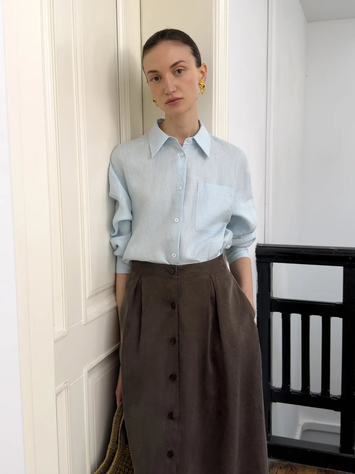 Model facing forward in light blue linen shirt with rolled sleeves, paired with brown pleated skirt