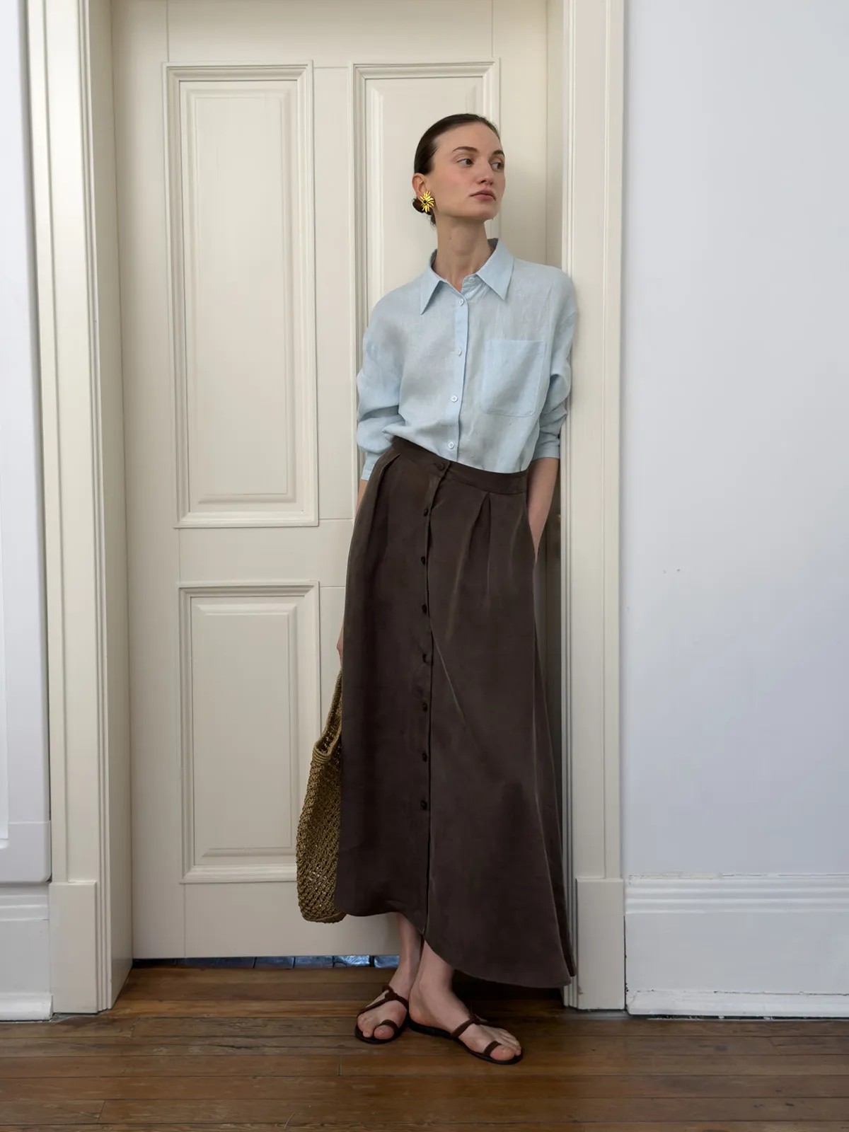 Model wearing light blue relaxed linen shirt with brown button-front midi skirt, standing beside white door