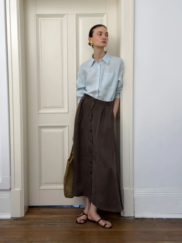 Model wearing light blue relaxed linen shirt with brown button-front midi skirt, standing beside white door