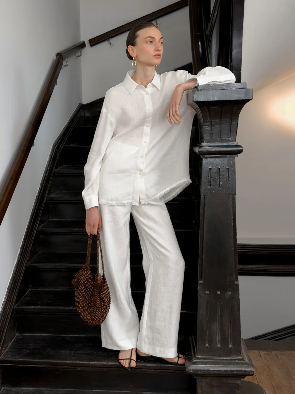 Model wearing white ELARA relaxed linen shirt and matching trousers on vintage wooden staircase, holding beaded bag