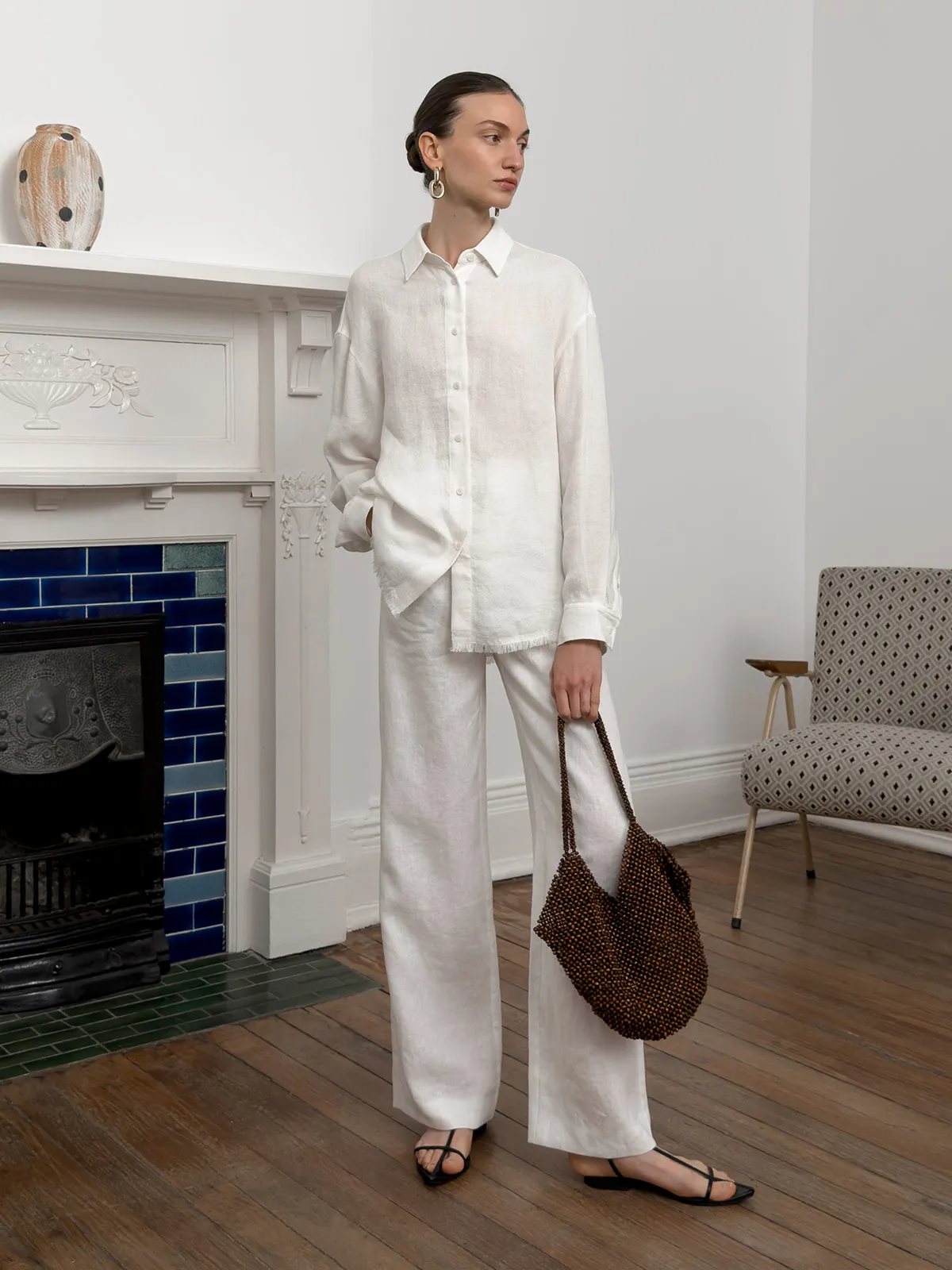 Model in ELARA shirt and trousers indoors, holding beaded bag beside patterned armchair and fireplace