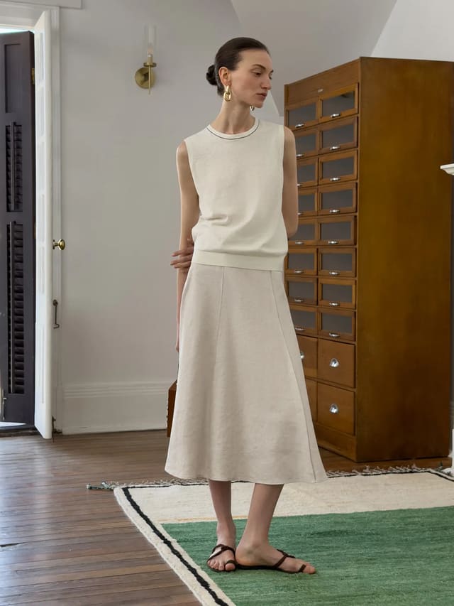 Model wearing NATASHA herringbone midi skirt with cream sleeveless knit top, standing indoors on green rug